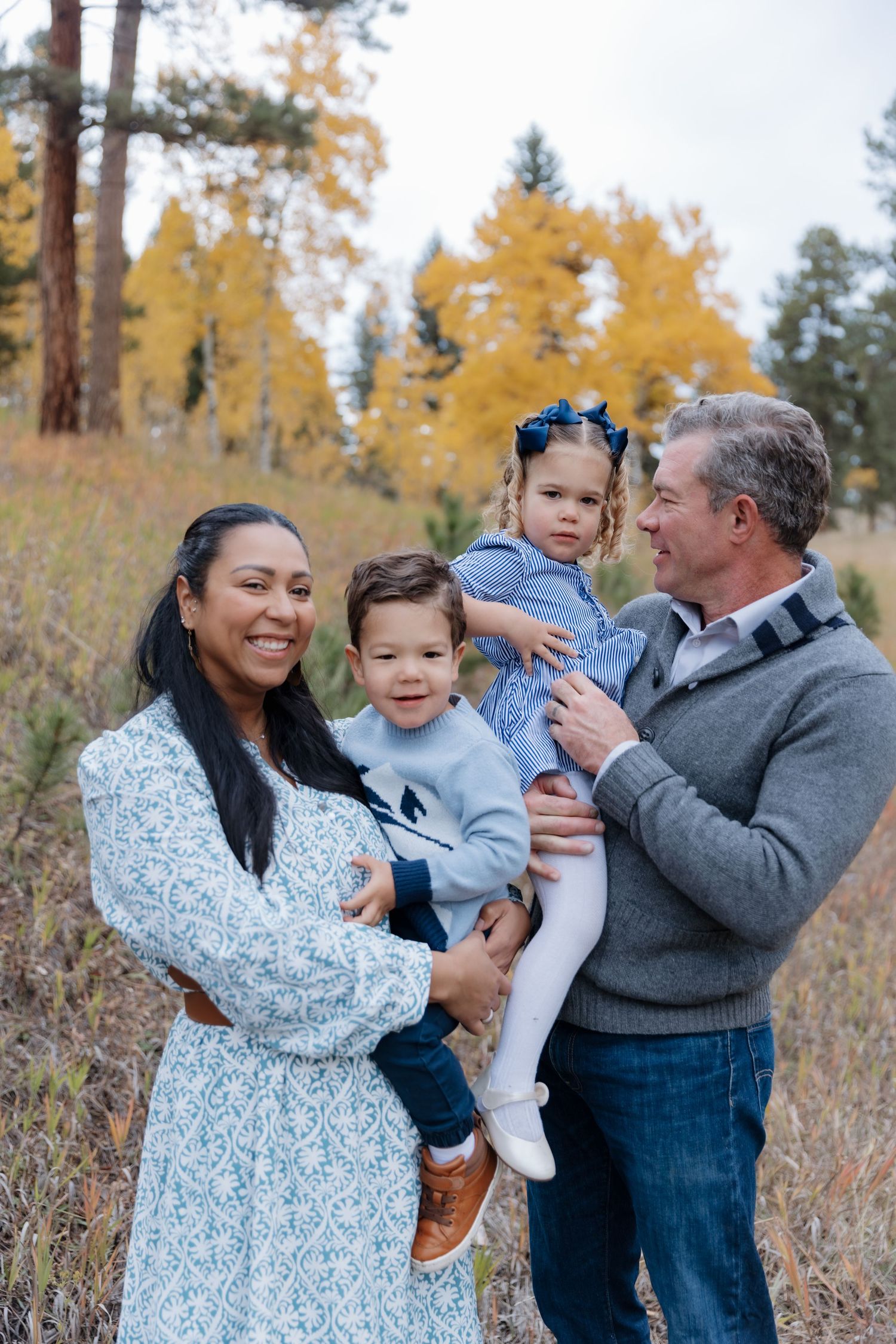 Family of four with two toddlers posing outdoors among golden fall foliage in a mountain meadow.