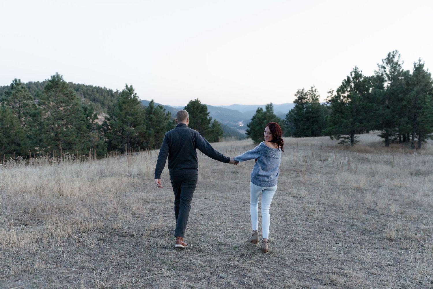 Couple holding hands while walking through a scenic mountain meadow at dusk.