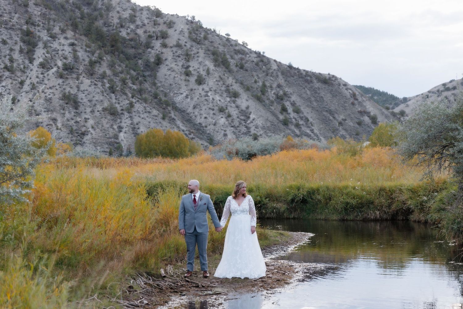 Bride and groom walk hand in hand along a scenic river with golden autumn foliage and rocky mountains behind.