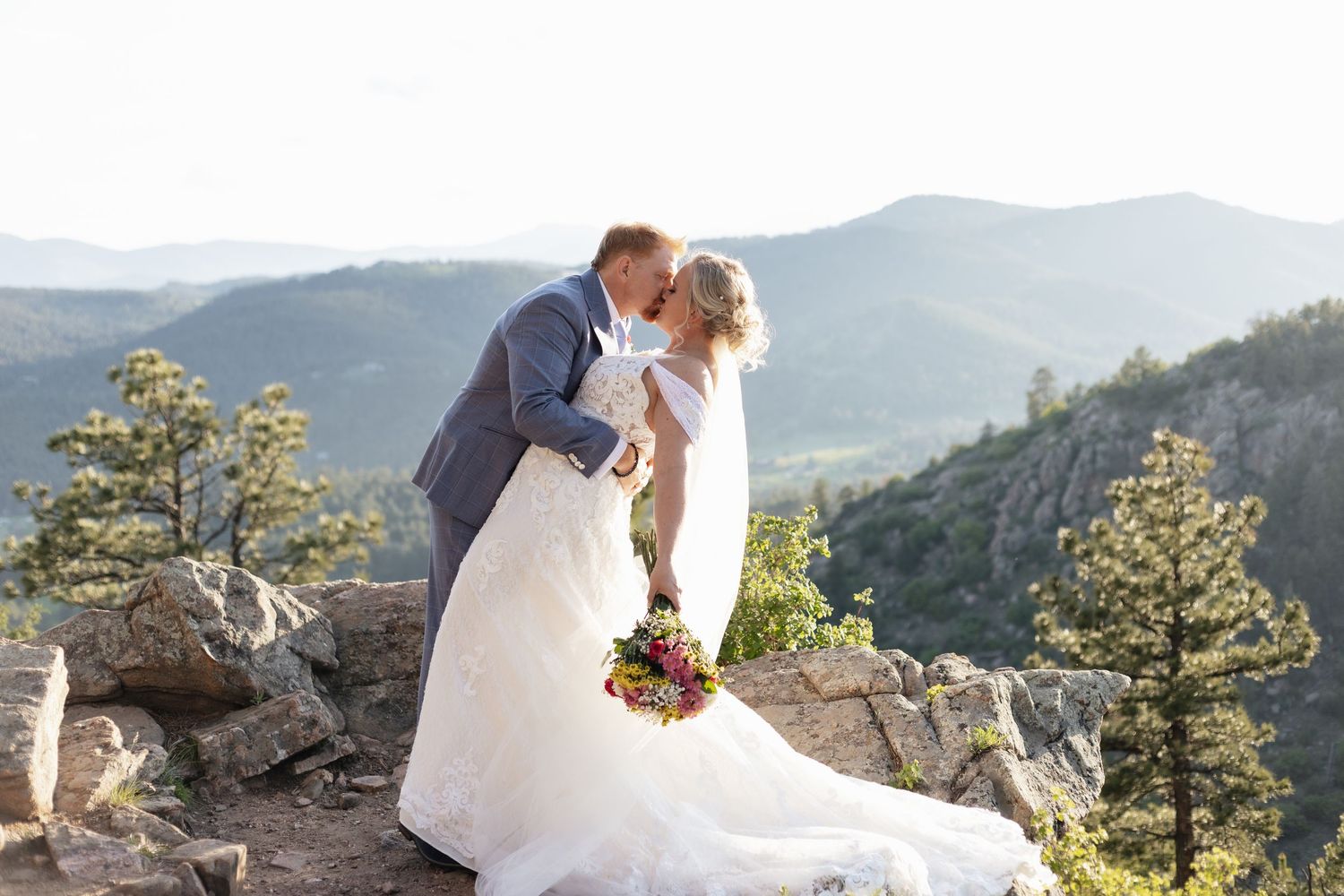 Bride and groom share kiss on rocky mountain overlook with scenic valley view.