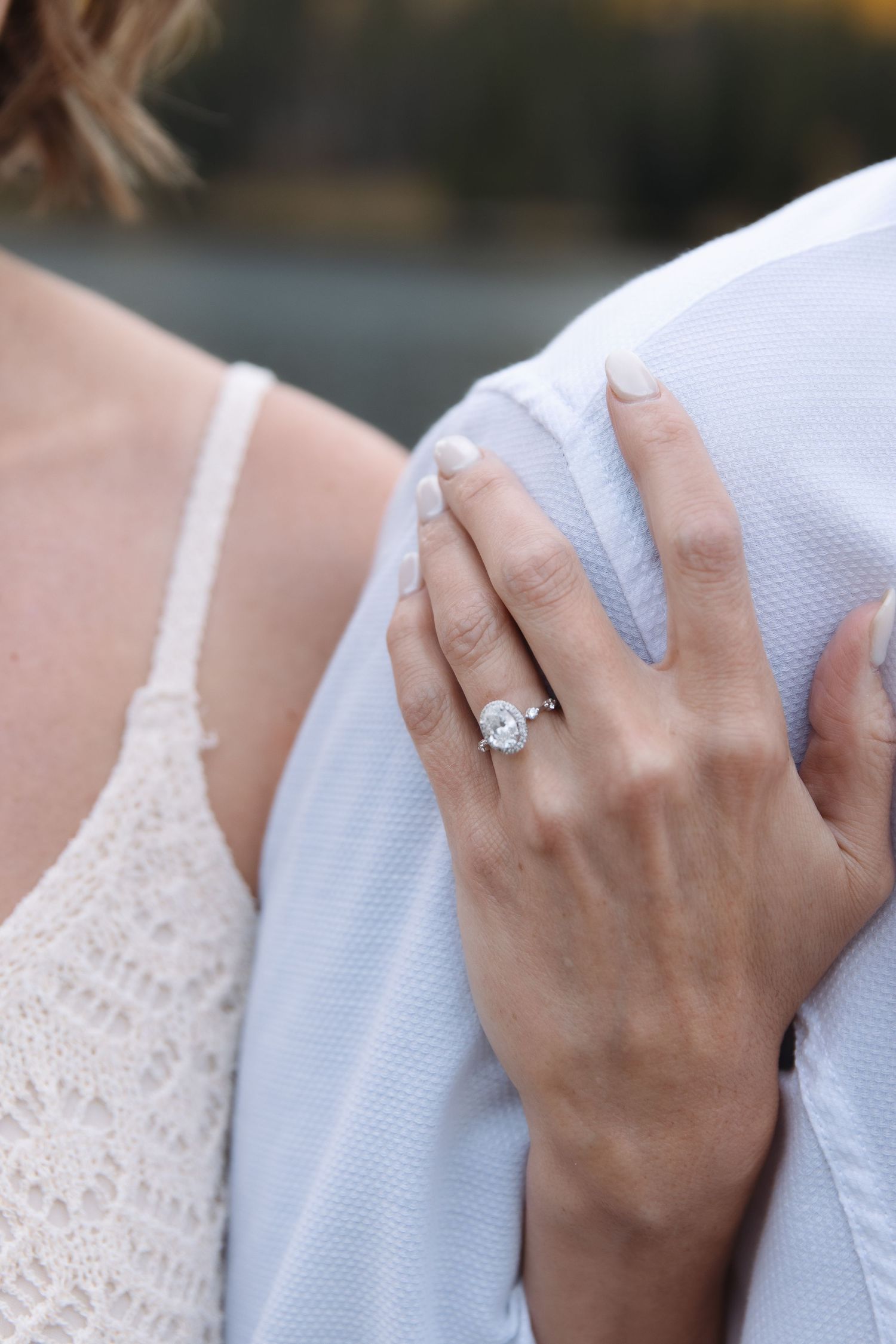 Close-up of an oval diamond solitaire engagement ring on womans hand resting on partners shoulder.