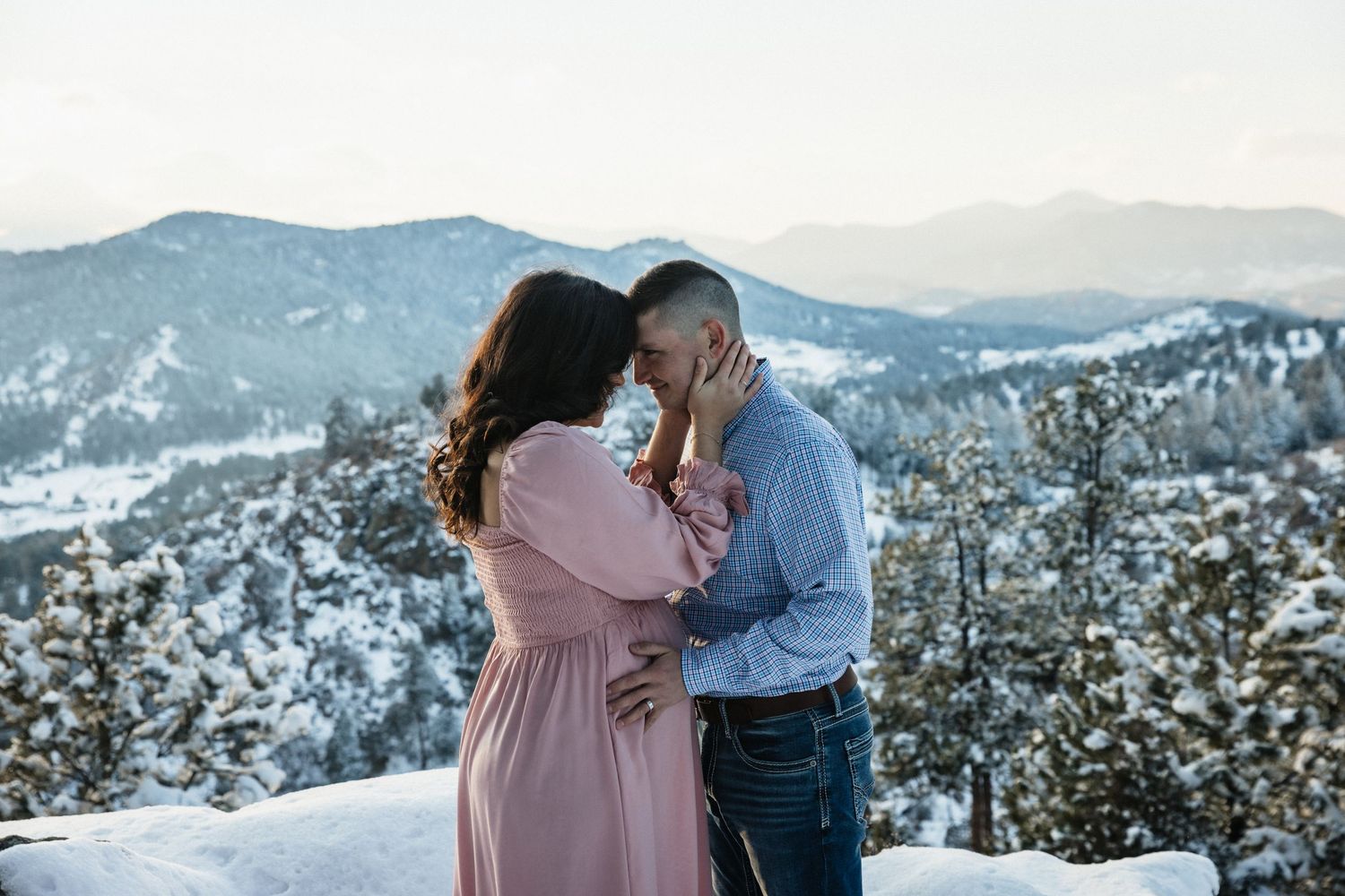 Couple sharing romantic kiss on snowy mountain overlook at sunset in pink and blue attire.