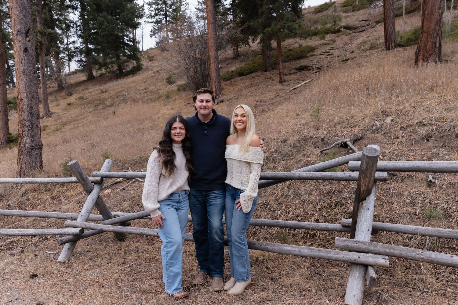 Three young adults smiling together leaning on a rustic wooden fence in an autumn mountain landscape.