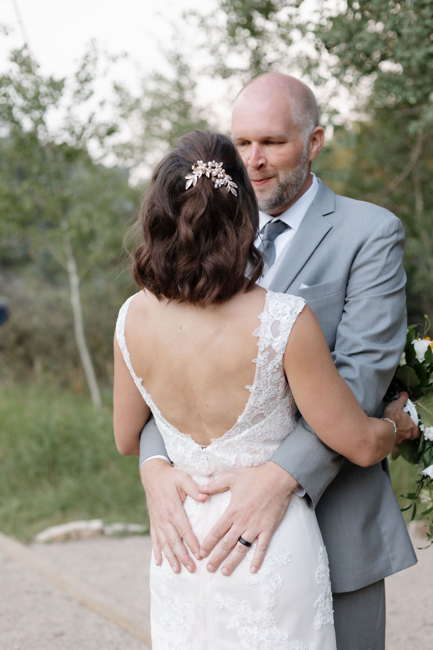 Groom in gray suit embracing bride in lace backless gown while holding bouquet outdoors.