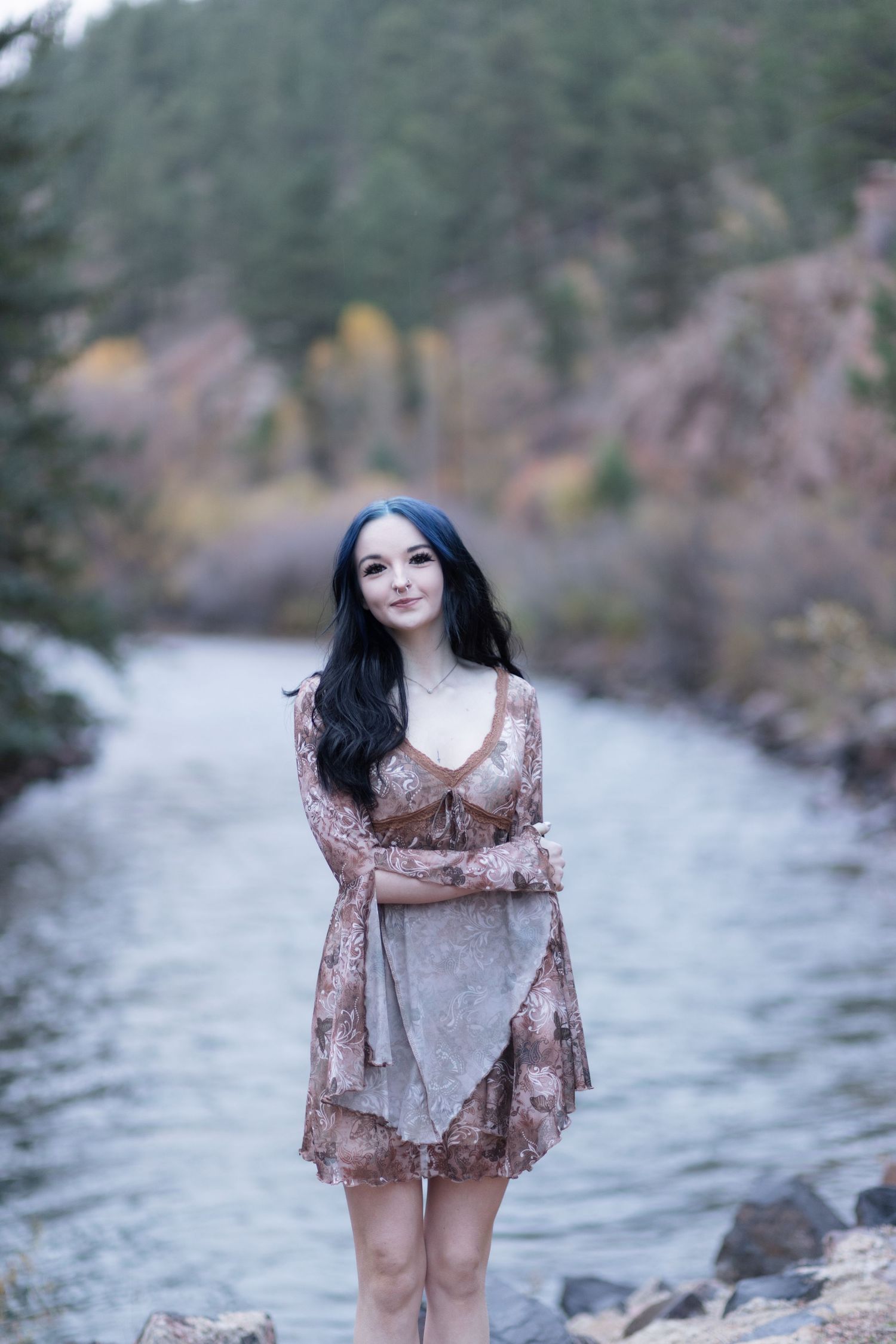 Young woman with dark hair in a floral dress standing by a serene mountain river in autumn.