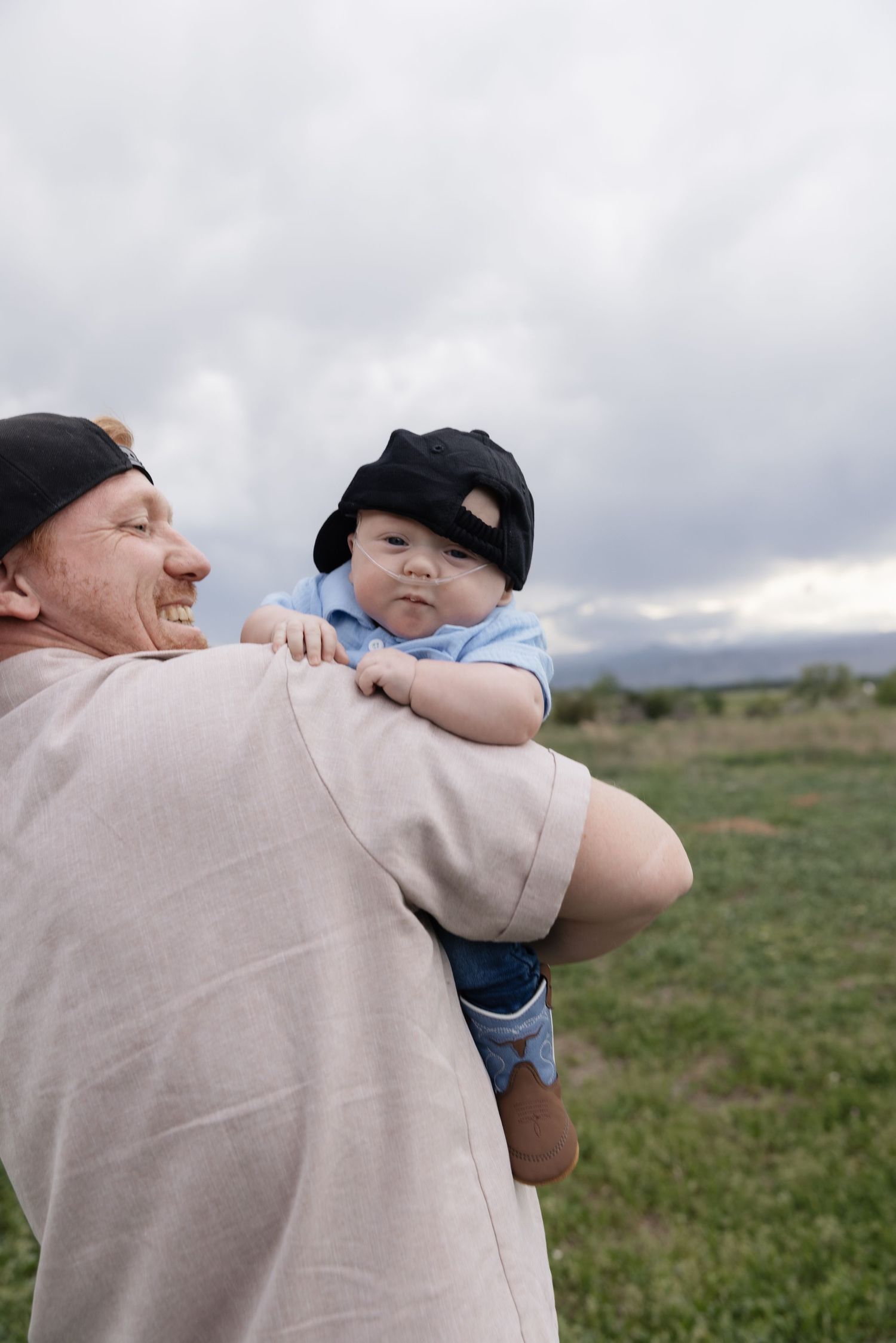 A smiling man holds a baby wearing a black cap outdoors in a green field under a cloudy sky.