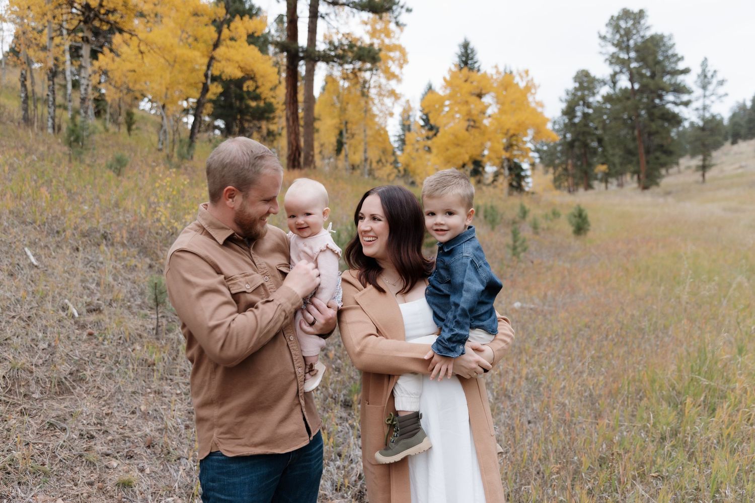 Young family with two toddlers posing in a golden autumn meadow surrounded by fall foliage.