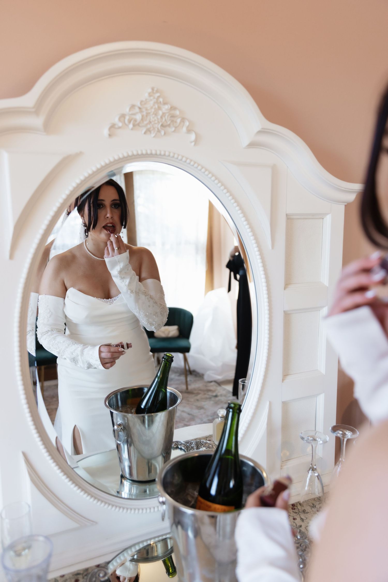 Bride in white gown applying lipstick while viewing reflection in ornate white mirror with champagne nearby.