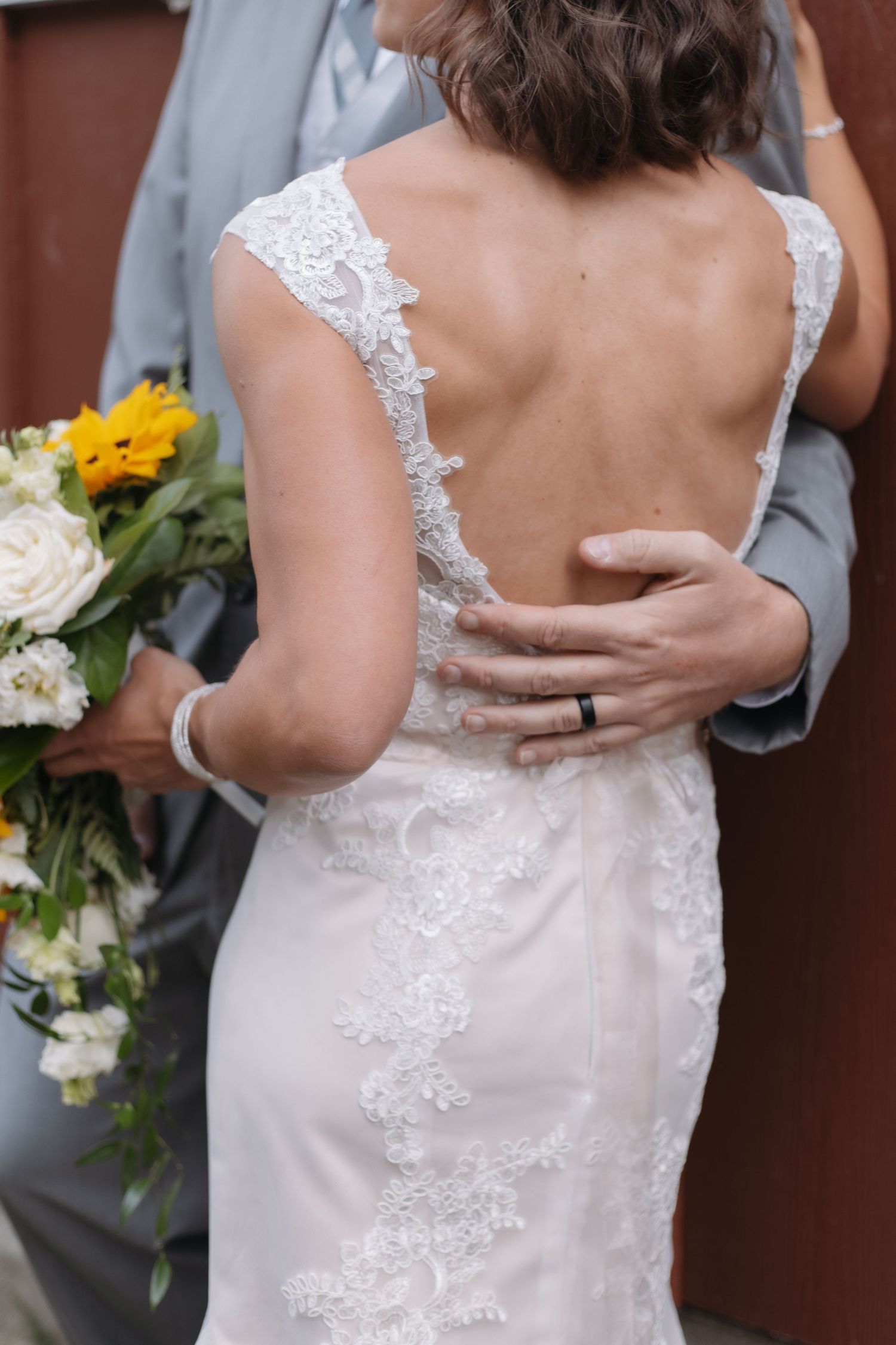 Bride in backless lace wedding gown held by groom holding sunflower bouquet.