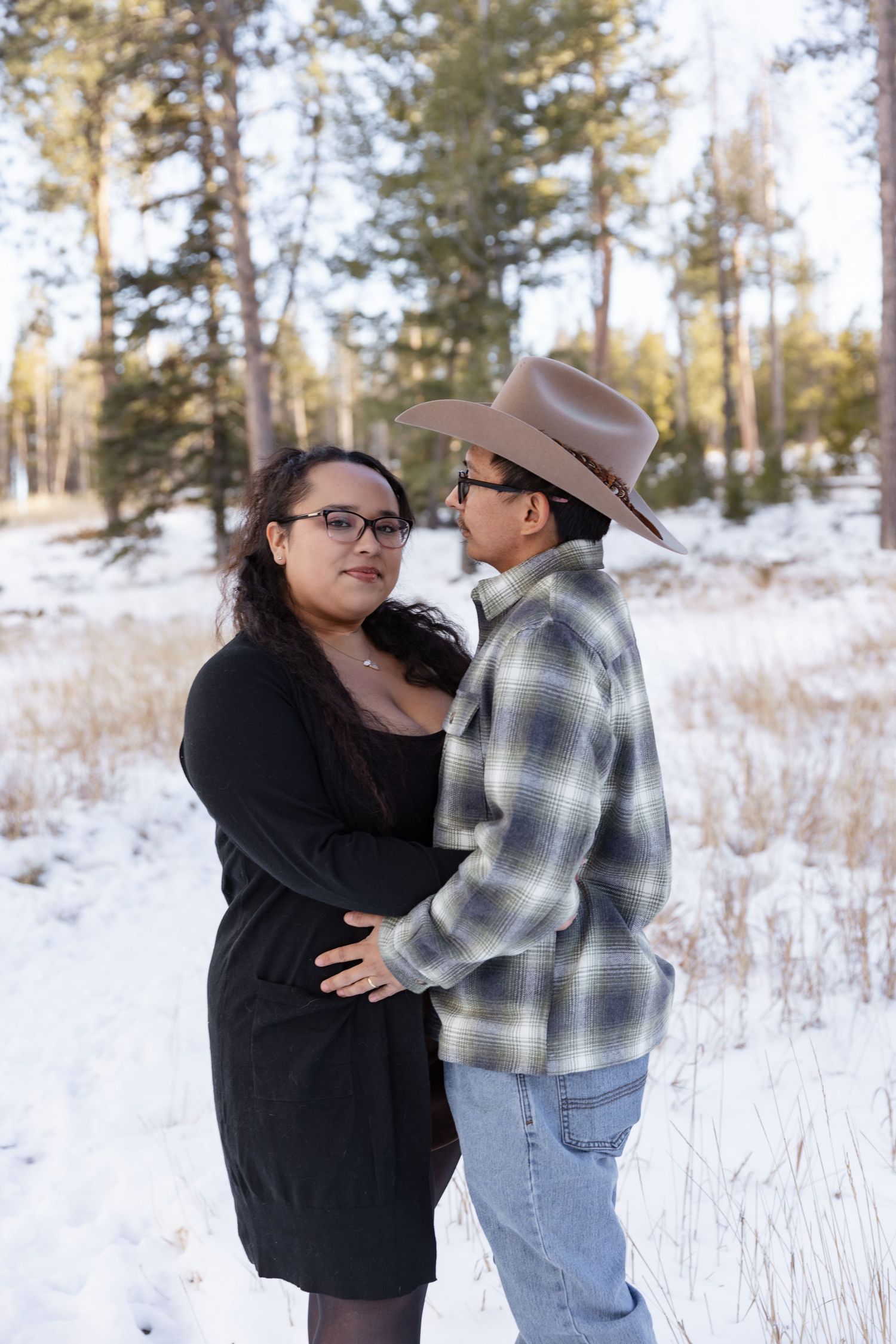 Couple embracing in snowy winter forest, woman in black dress and man in plaid shirt and cowboy hat.
