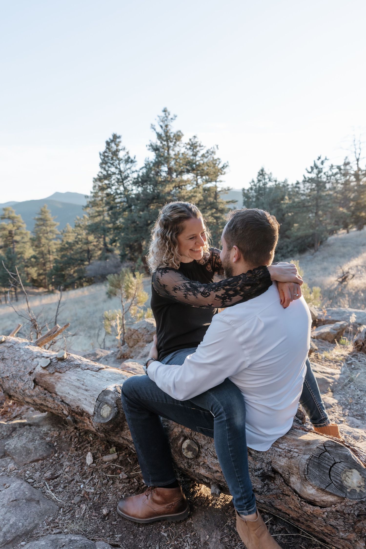 Couple sitting on a fallen log in a pine forest, sharing a tender moment outdoors.
