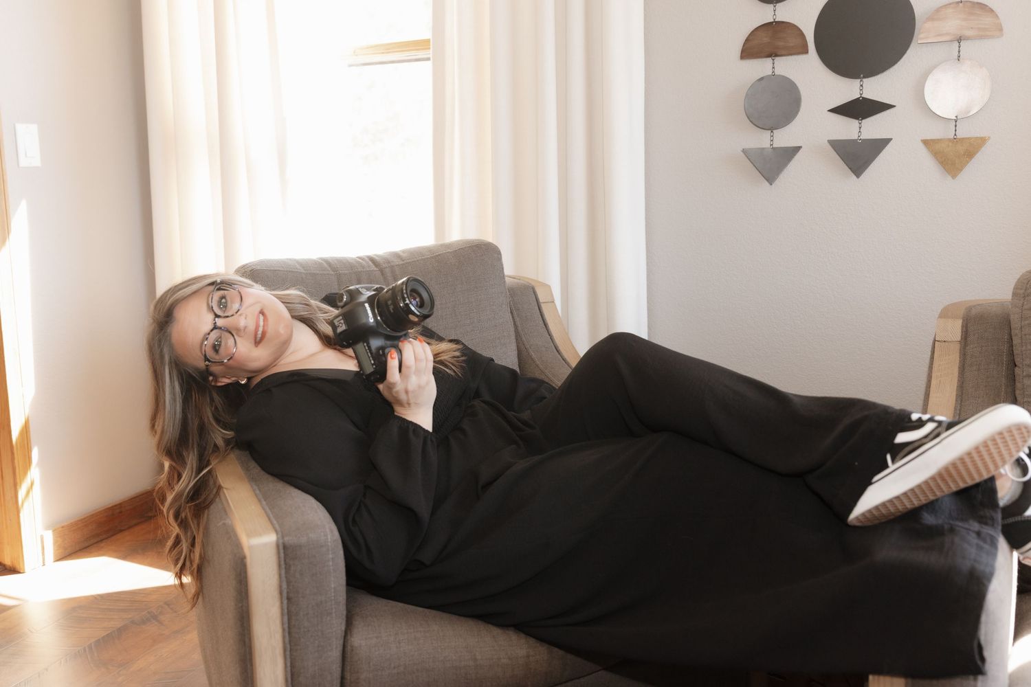 Woman relaxes on gray sofa holding camera in bright modern room.