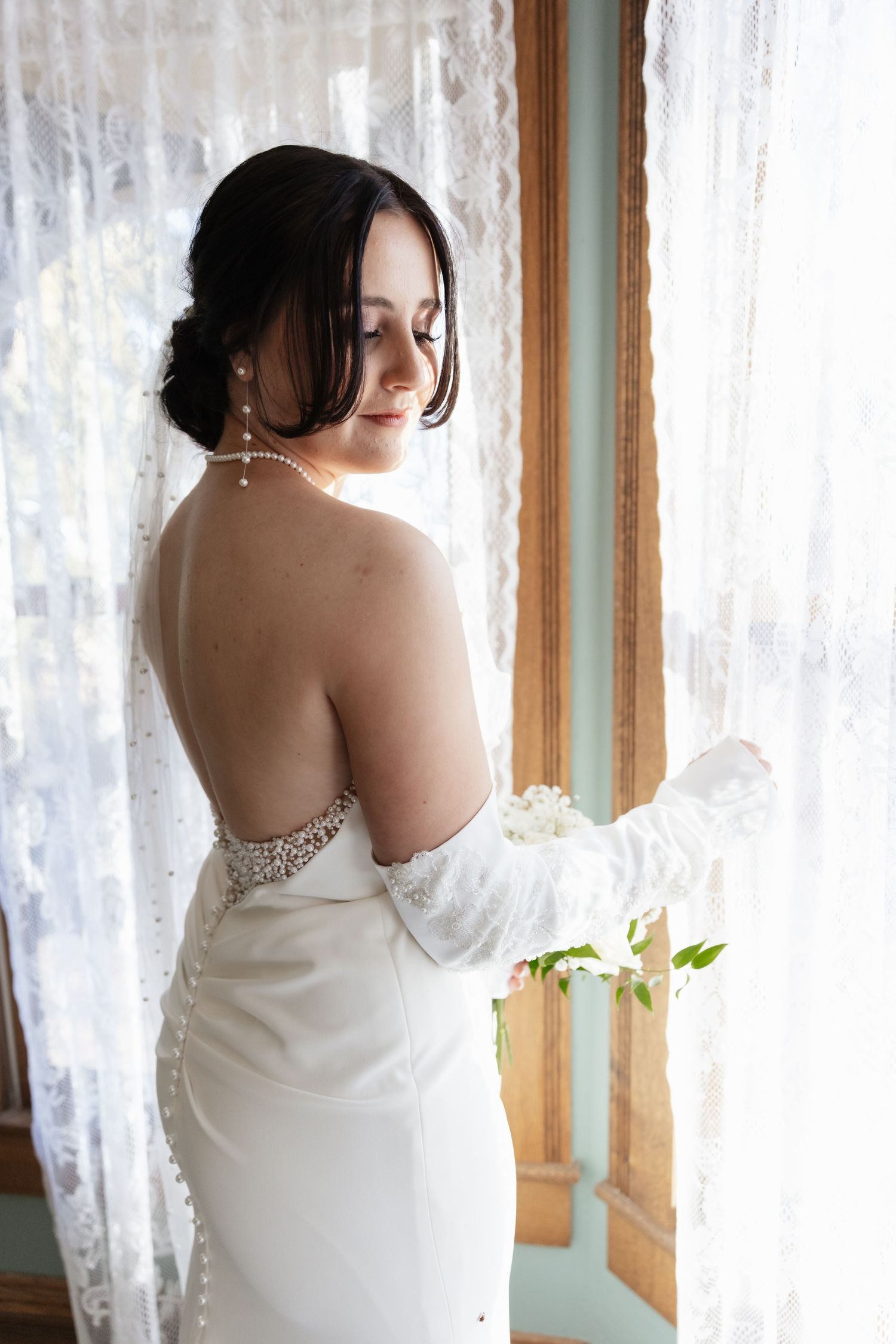 Elegant bride in backless white gown holding flowers, standing by lace curtained window with pearl jewelry.