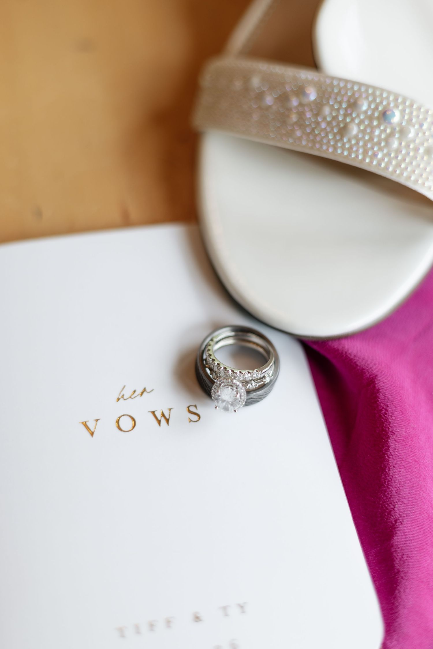 Wedding rings stacked on a vow book beside a pearl-embellished bridal heel and pink fabric.