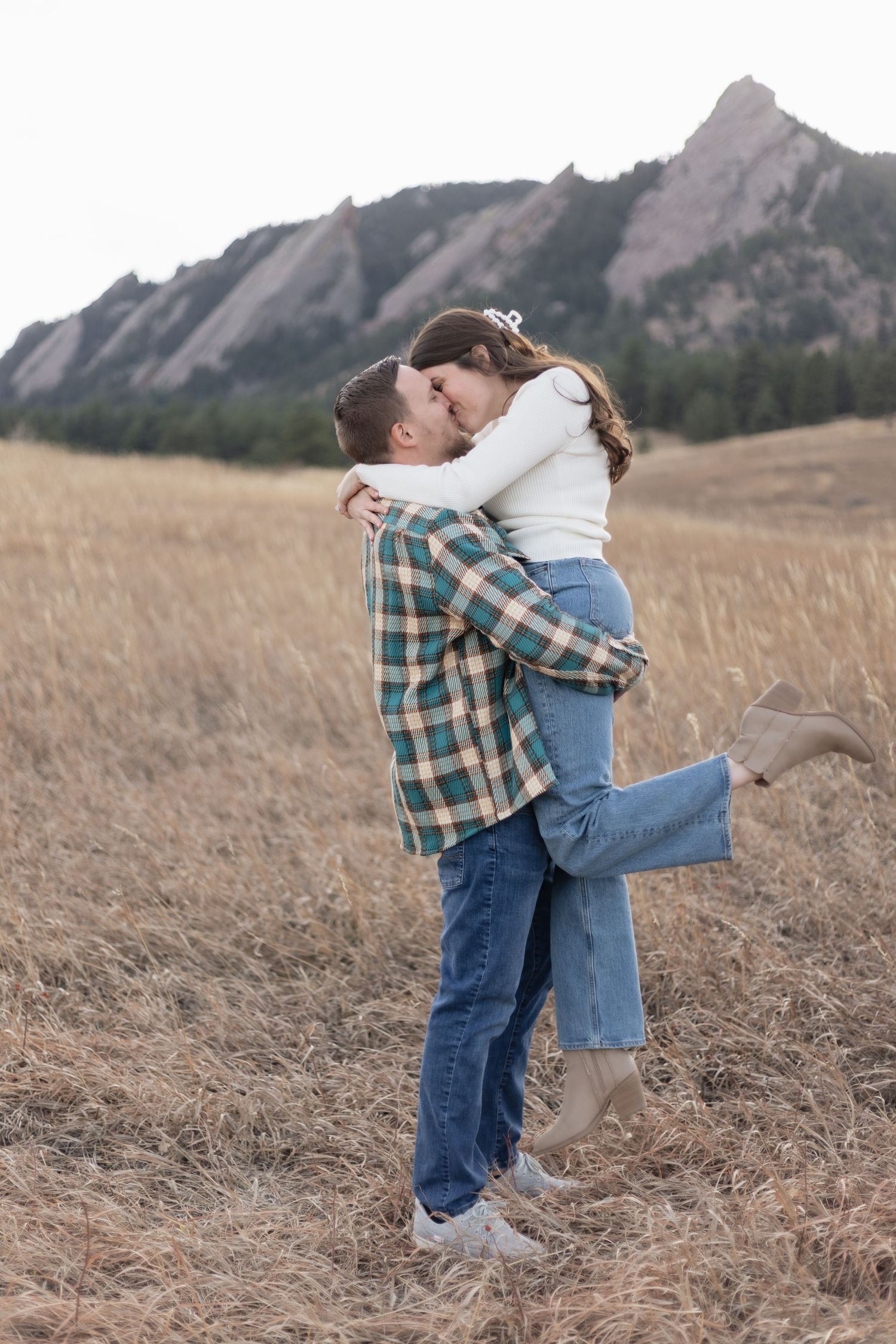 Man lifts woman for kiss in golden field with rocky mountains. Flatirons