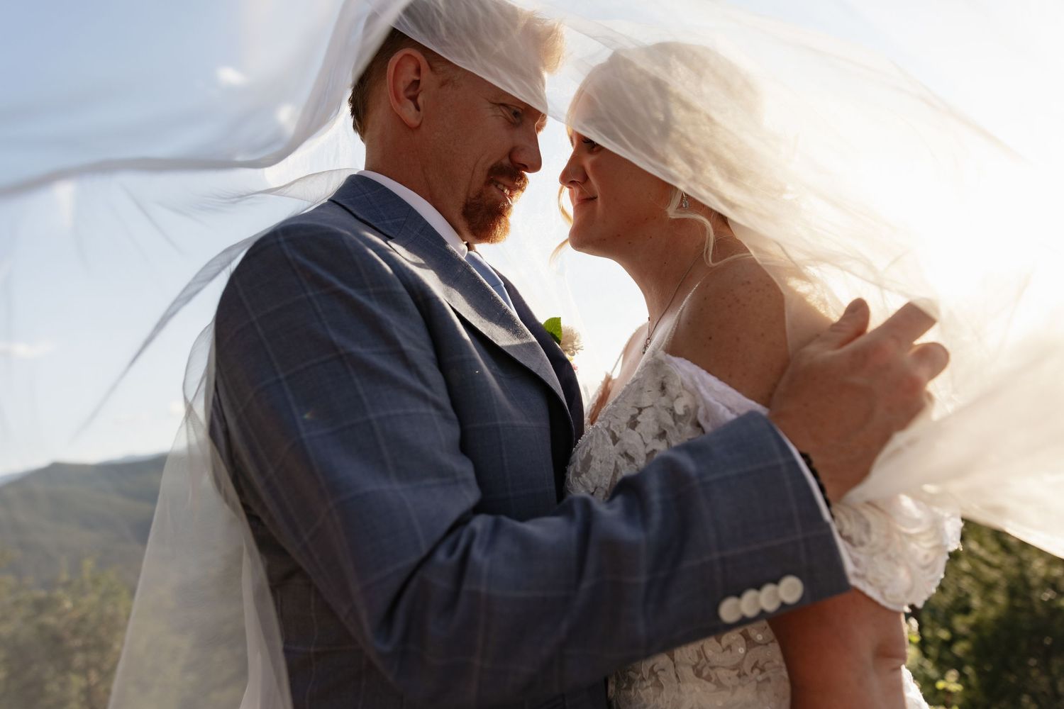 Bride and groom share intimate moment under flowing veil with mountain backdrop.