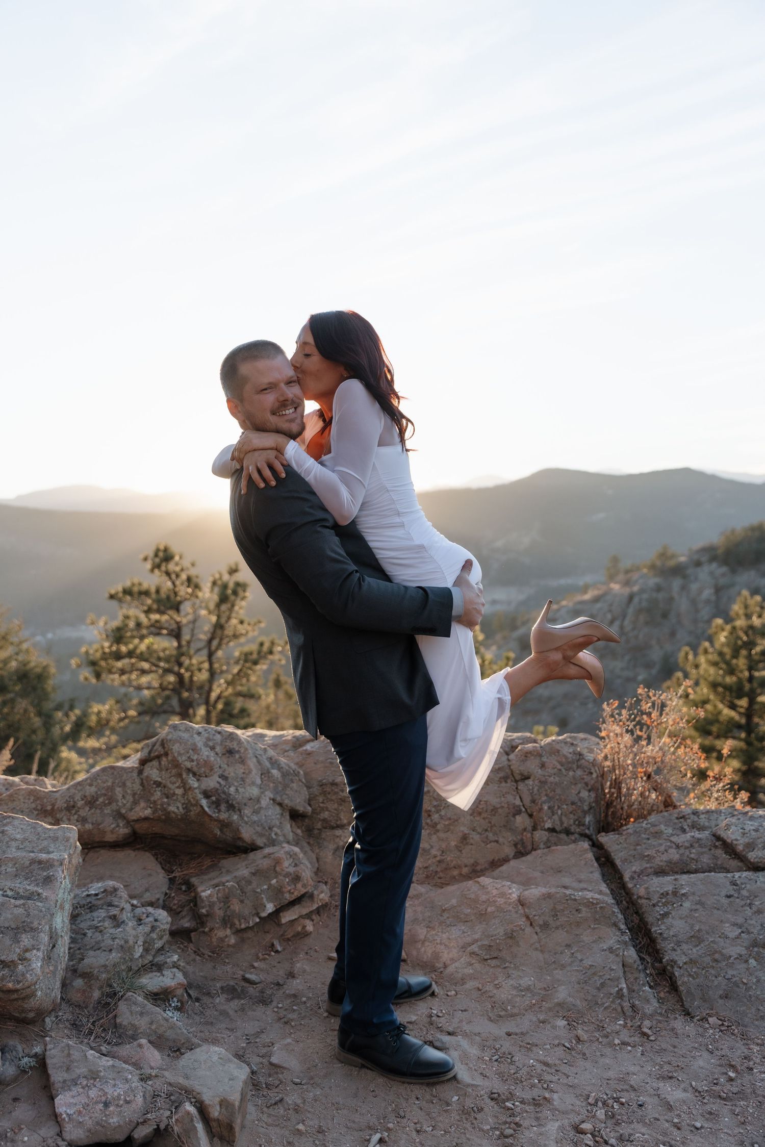 Groom lifting bride for a kiss on rocky overlook with stunning golden sunset mountain backdrop.