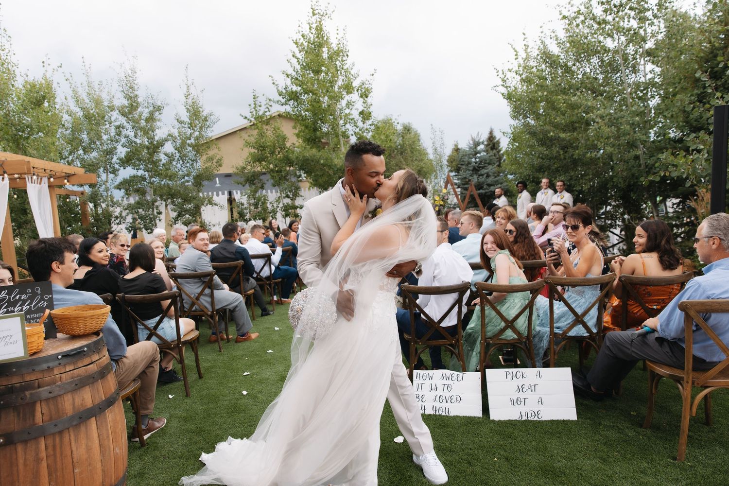 Groom dips bride in romantic outdoor wedding reception kiss surrounded by seated guests and rustic barrel decorations.