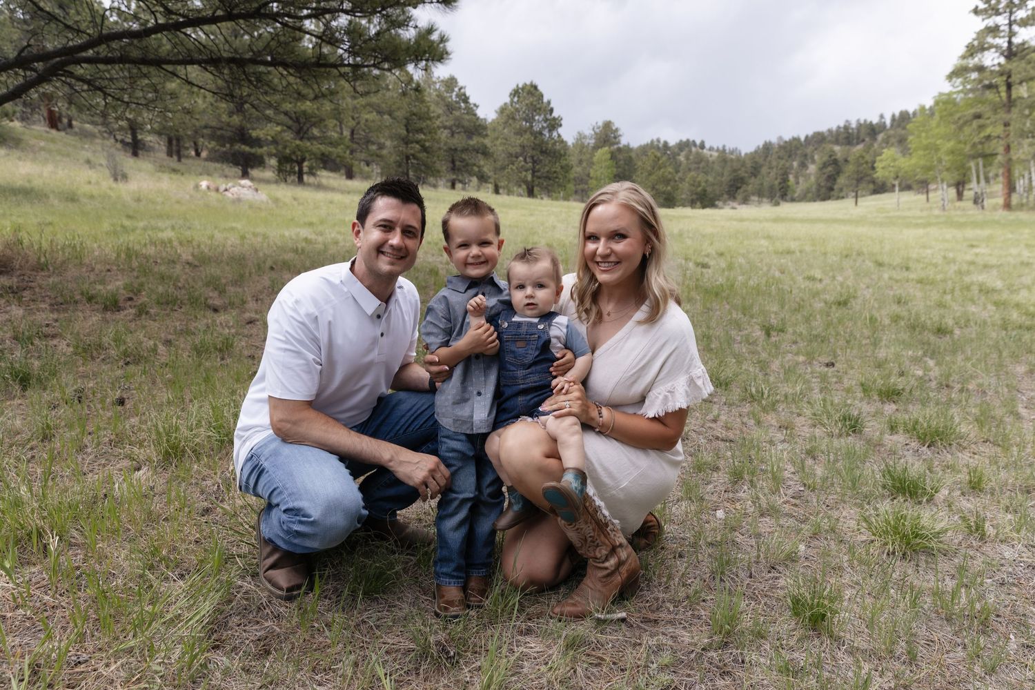 Family of four posing outdoors in a grassy meadow surrounded by trees, parents crouching with two young boys between them.