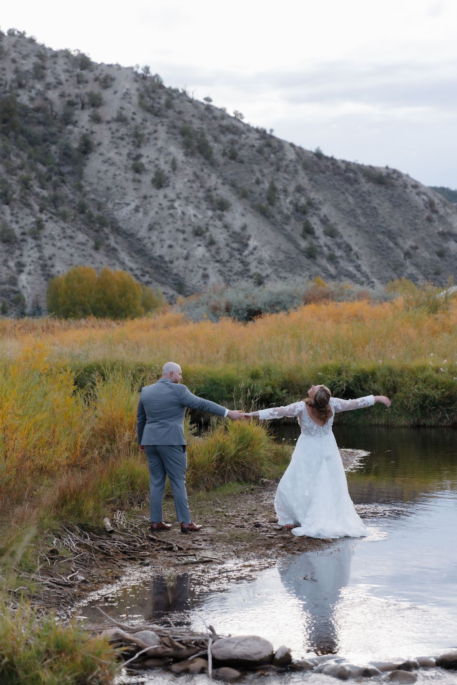 Bride and groom holding hands near a mountain stream surrounded by autumn foliage.
