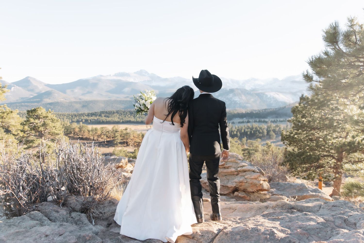 Bride and groom in western attire admire stunning mountain landscape views from rocky overlook at their elopement.