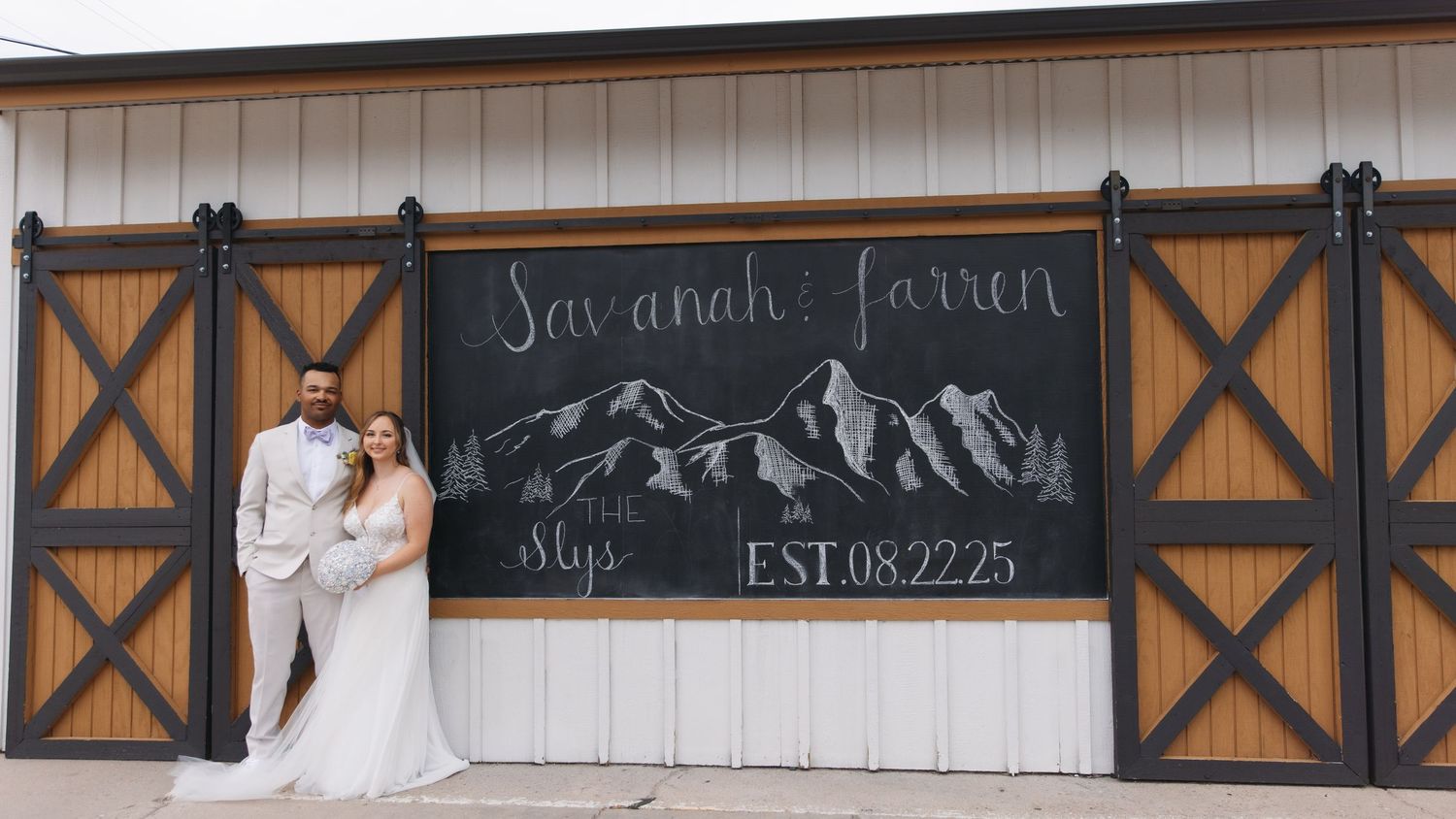 Bride and groom pose before rustic barn wedding sign outdoors.