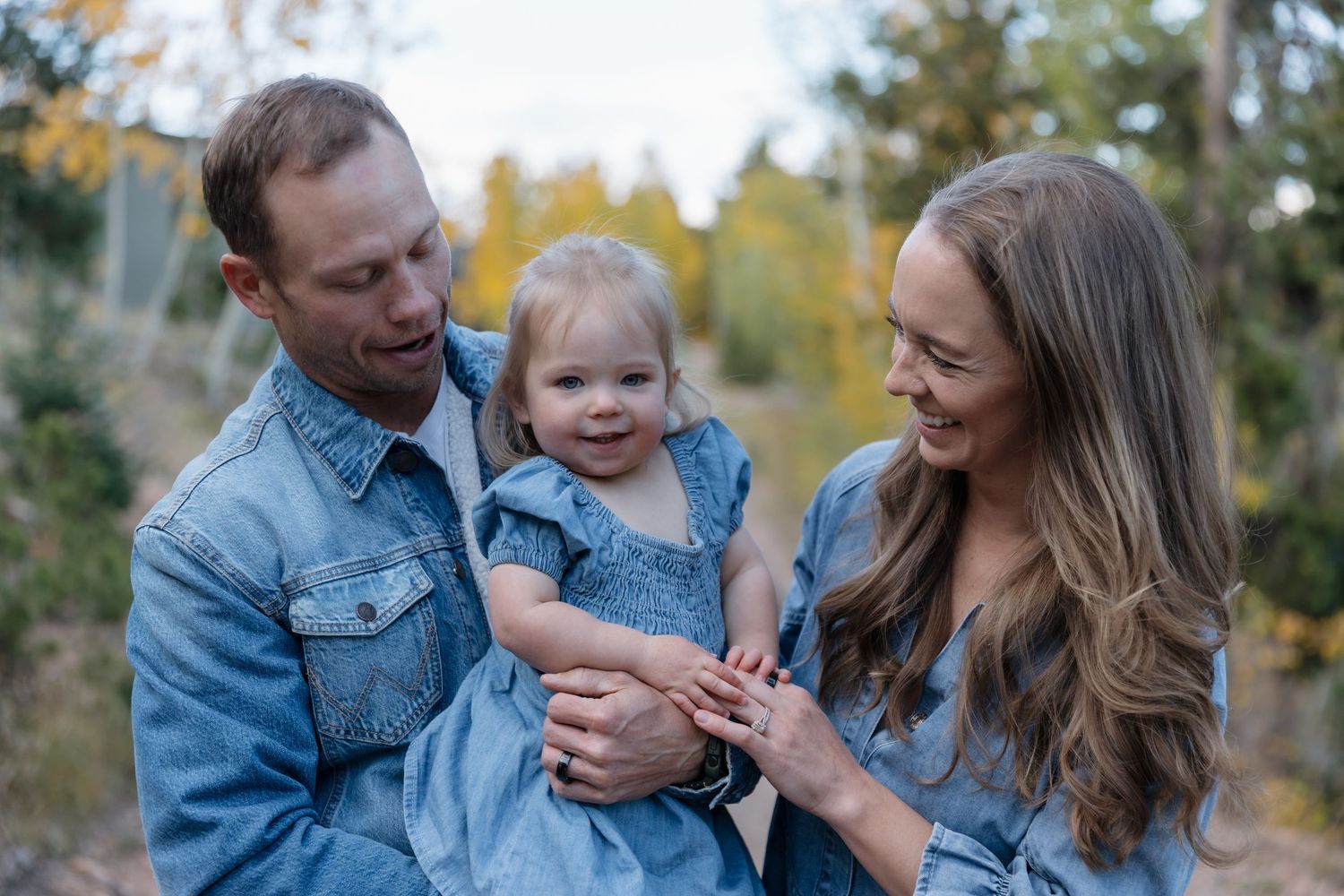 Happy family of three in denim outfits laughing together outdoors among fall foliage.