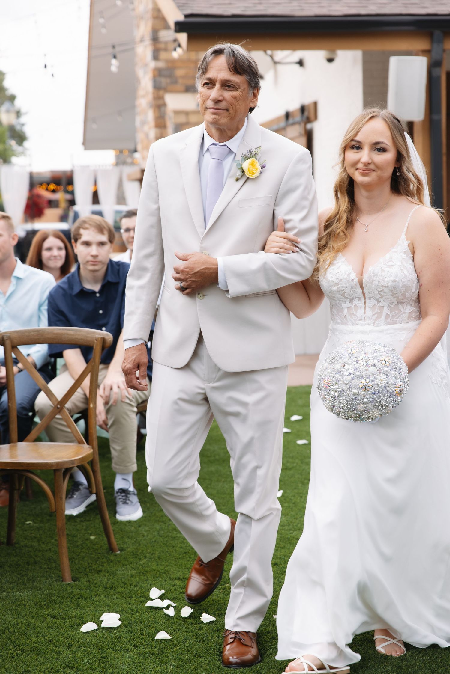 Father walks blonde bride in lace gown down outdoor wedding aisle.