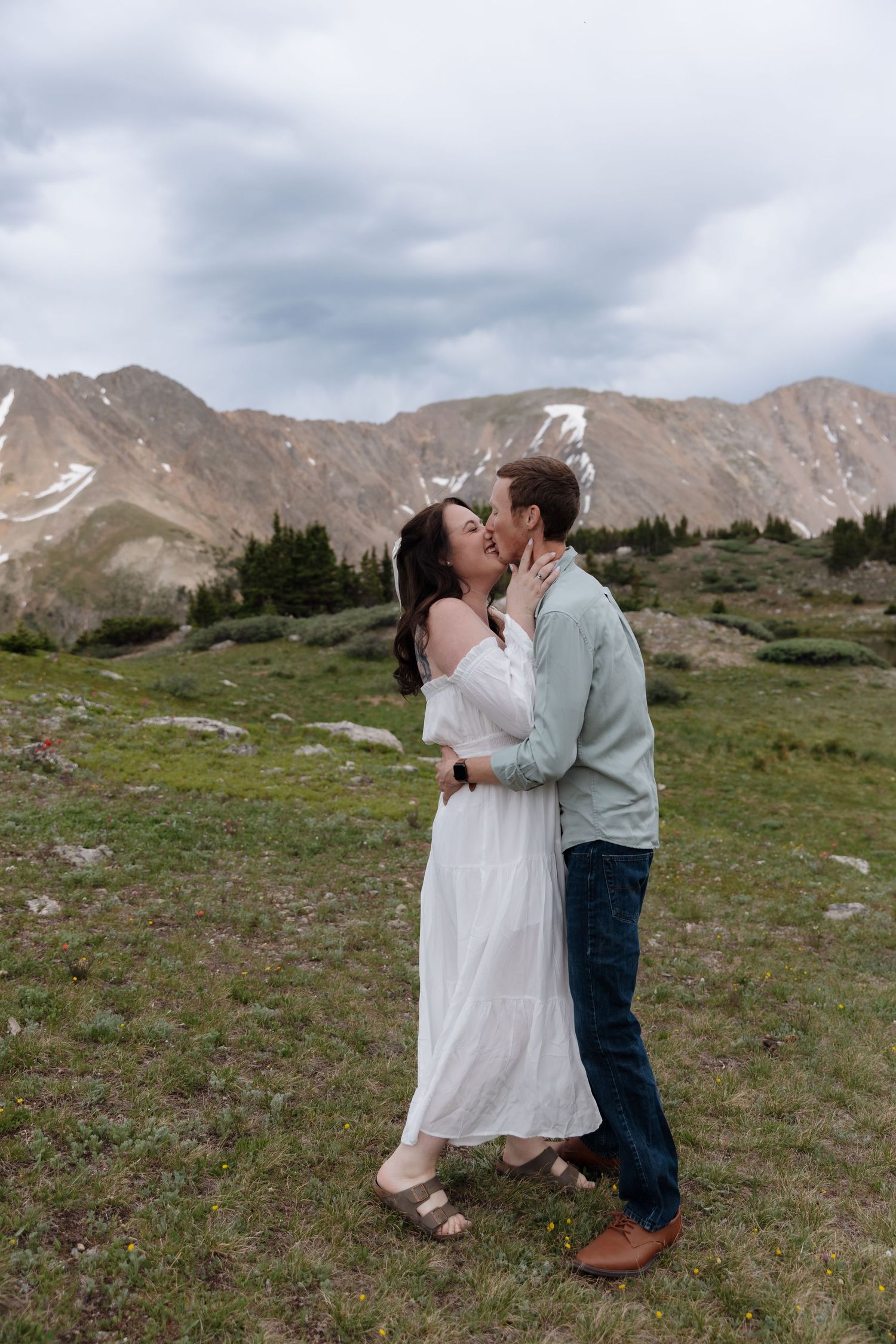 Couple sharing a romantic kiss in a green alpine meadow with snowy mountain backdrop.