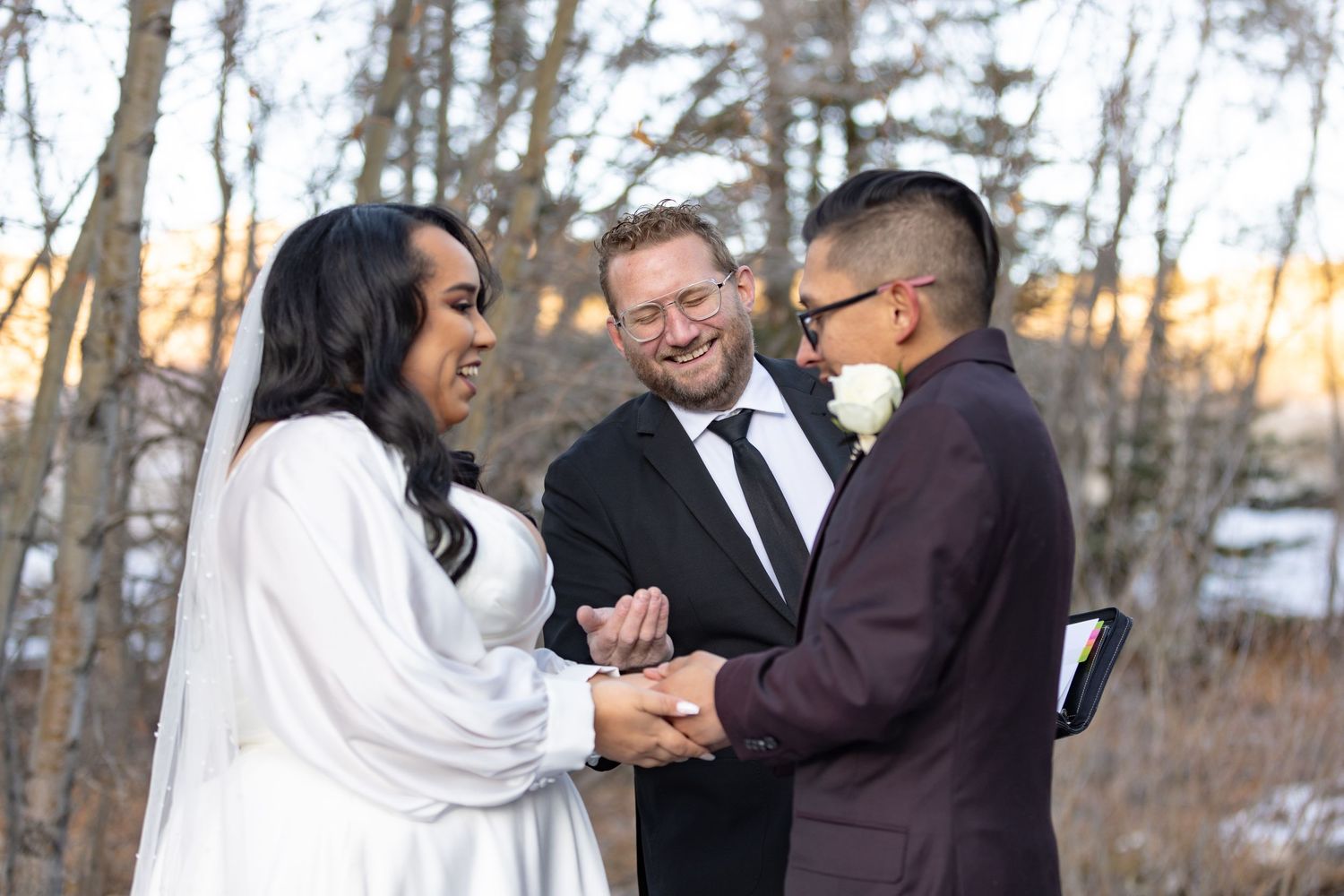 Same-sex couple exchanges vows while holding hands with smiling officiant during outdoor winter wedding ceremony.