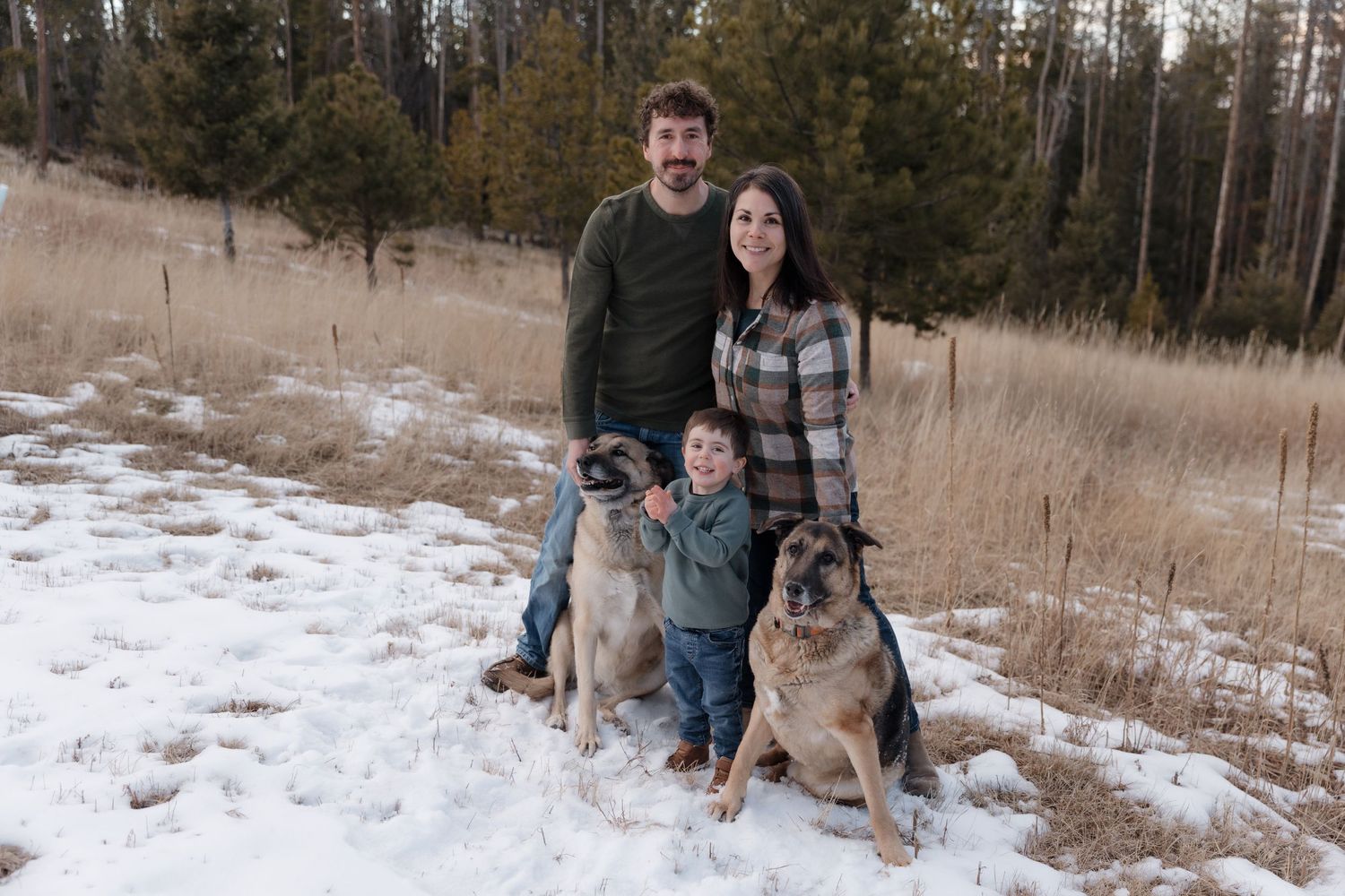 Family with young child and two German Shepherds posing in a snowy pine forest.