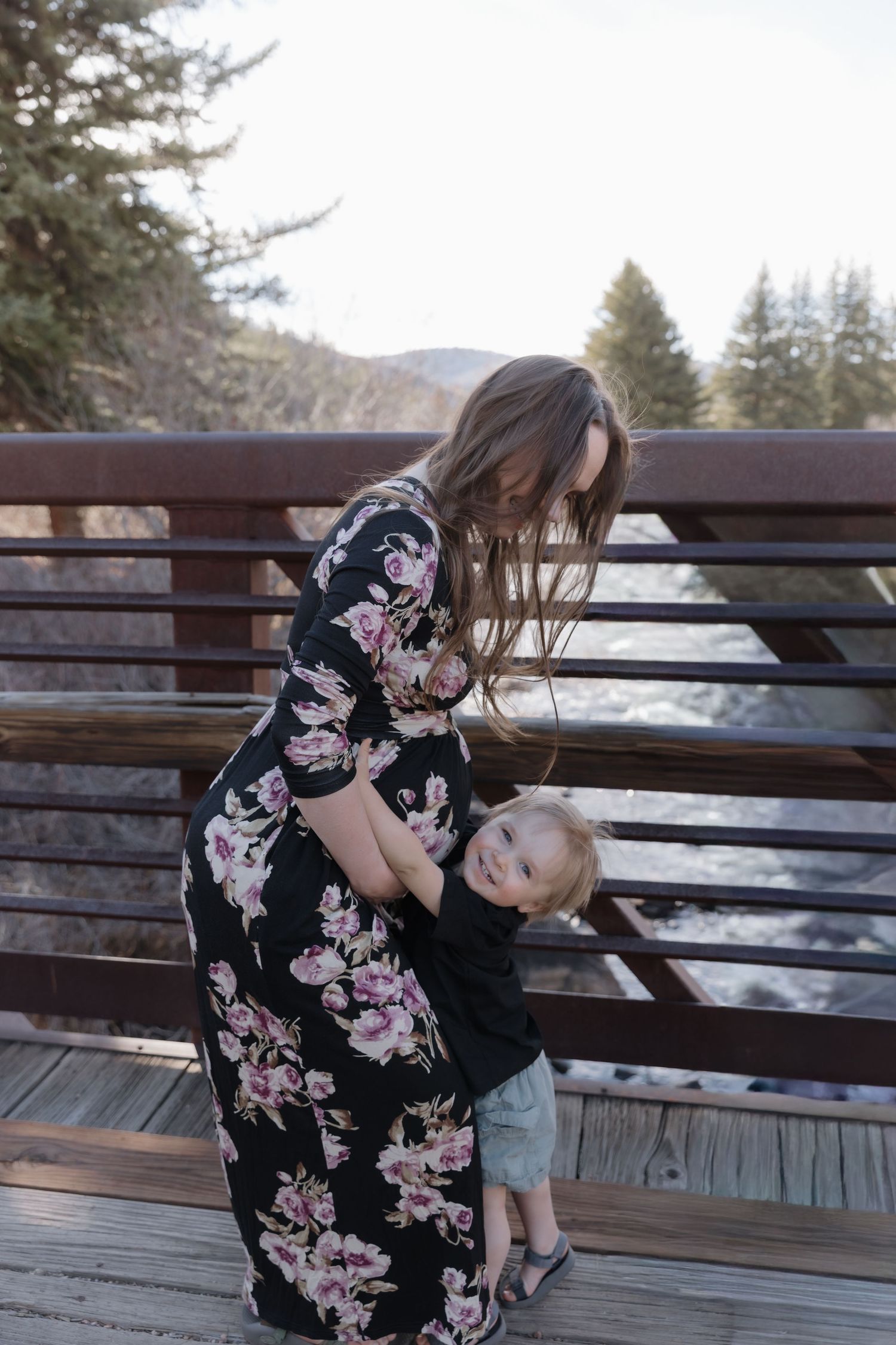 Mother in floral dress laughing with toddler on a wooden bridge near a mountain stream.