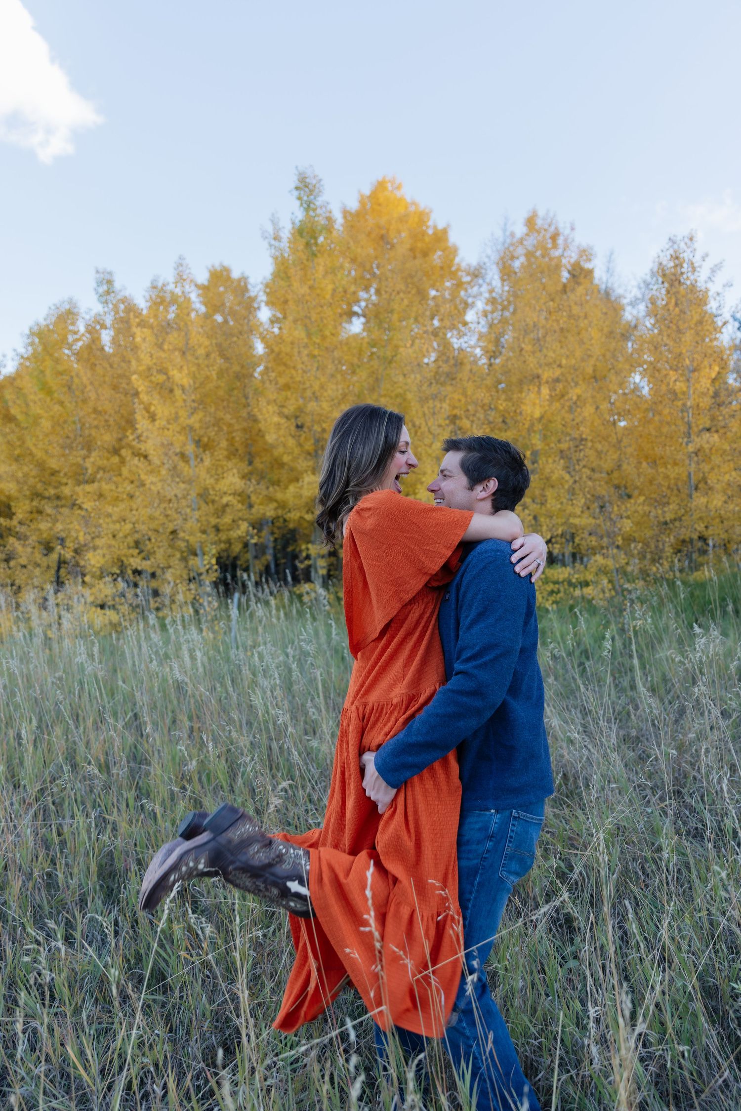 Couple embracing in a green meadow with vibrant yellow autumn trees under a clear blue sky.