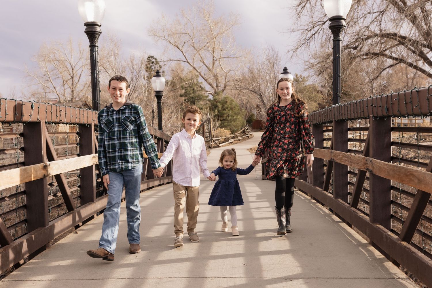 Family of four holding hands walking across a wooden bridge in a park during winter.
