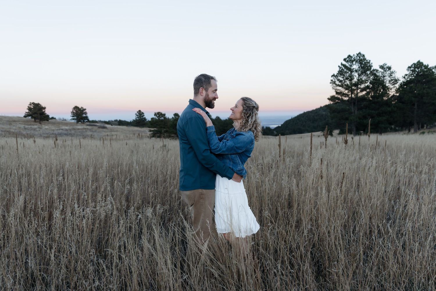 Romantic couple embracing in a golden field at dusk with pine trees in background.