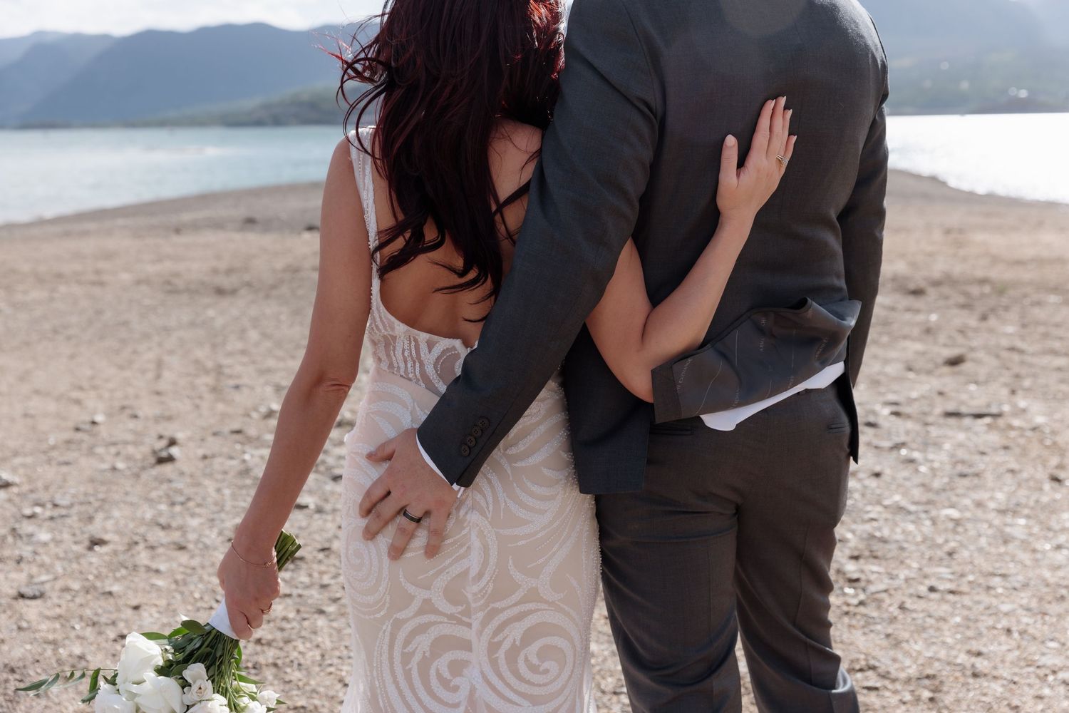 Bride and groom embracing on a beach, bride holding white bouquet in elegant lace wedding dress.