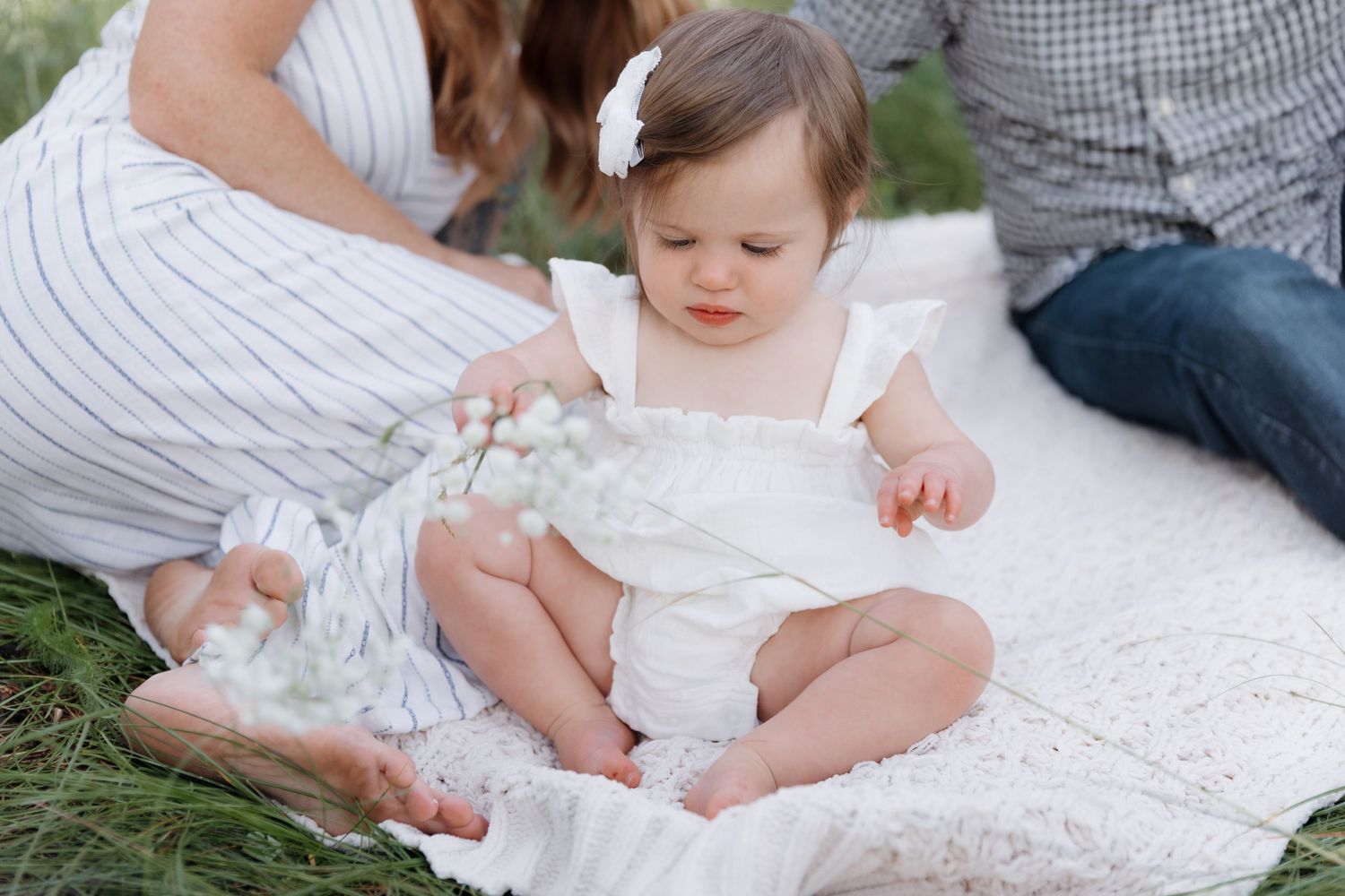 Baby girl in white romper sitting on blanket outdoors examining delicate white flowers.