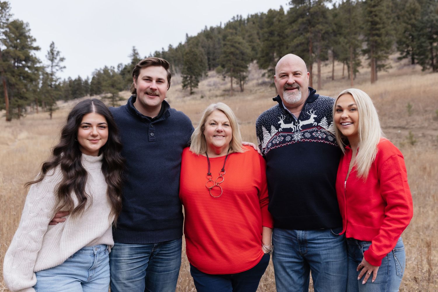 Family of five smiling outdoors in a mountain meadow wearing holiday sweaters and festive attire.
