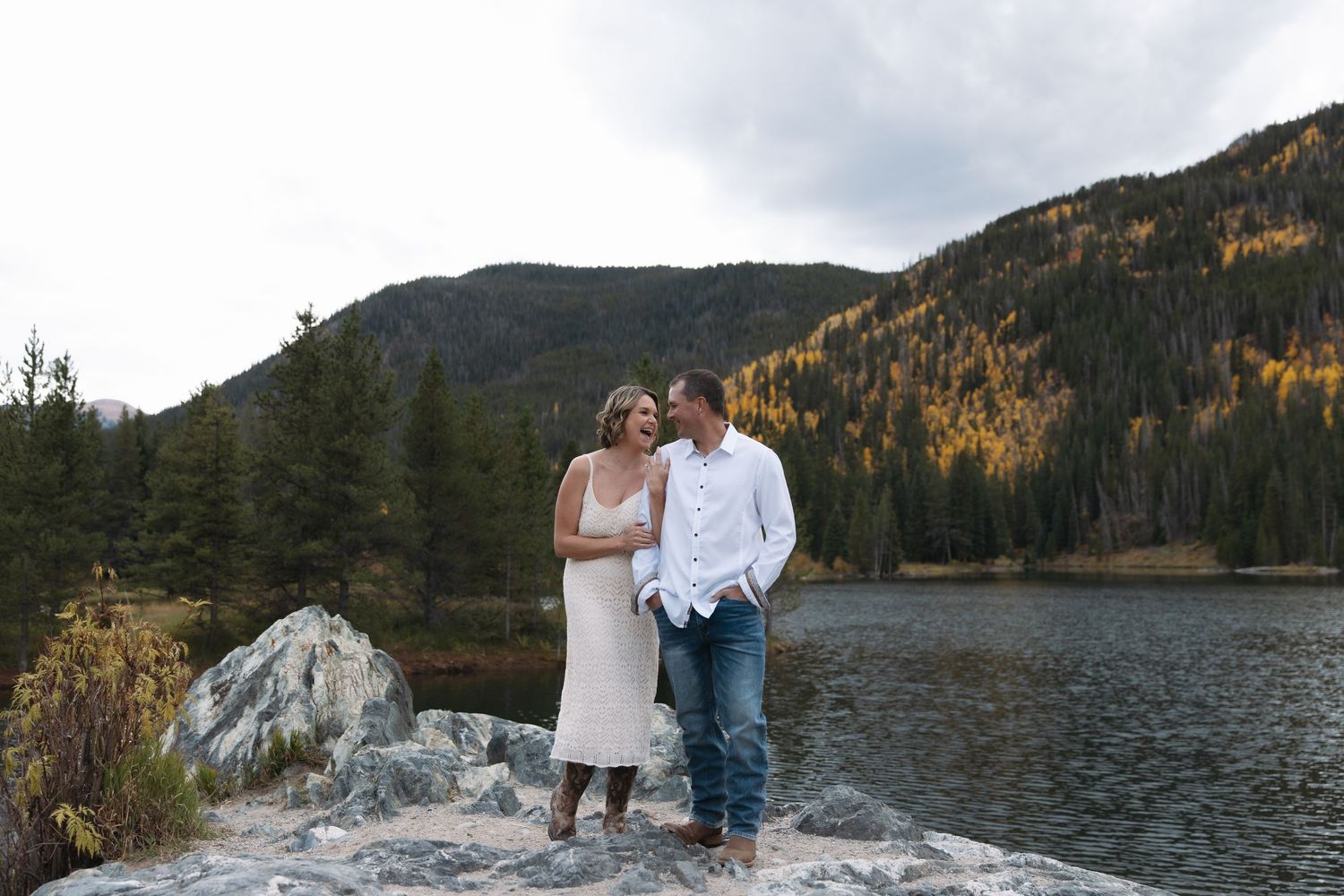 Couple standing on rocks by a mountain lake surrounded by autumn trees in Colorado.