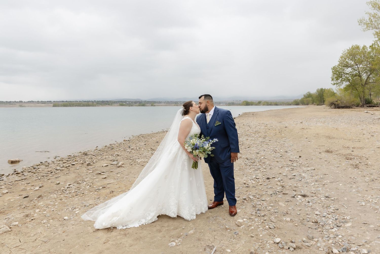 Bride in flowing white gown and groom in navy suit kissing by a lakeside shore.