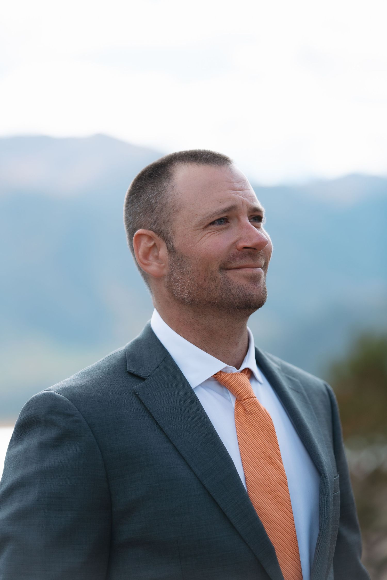 Handsome groom in gray suit and orange tie smiling with mountain landscape behind him.