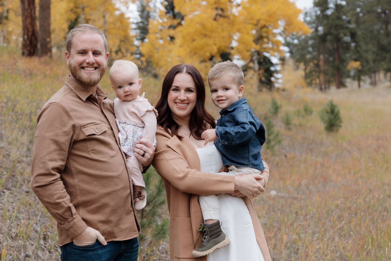 Happy family of four with two young children posing in a golden autumn meadow with yellow trees.