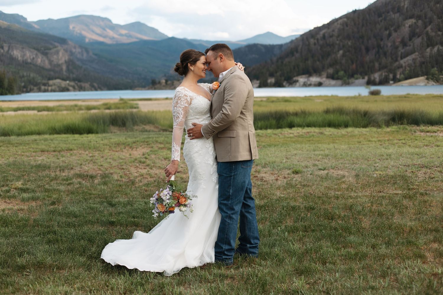 Bride and groom embrace in a mountain meadow with a scenic lake and mountain backdrop at their wedding.