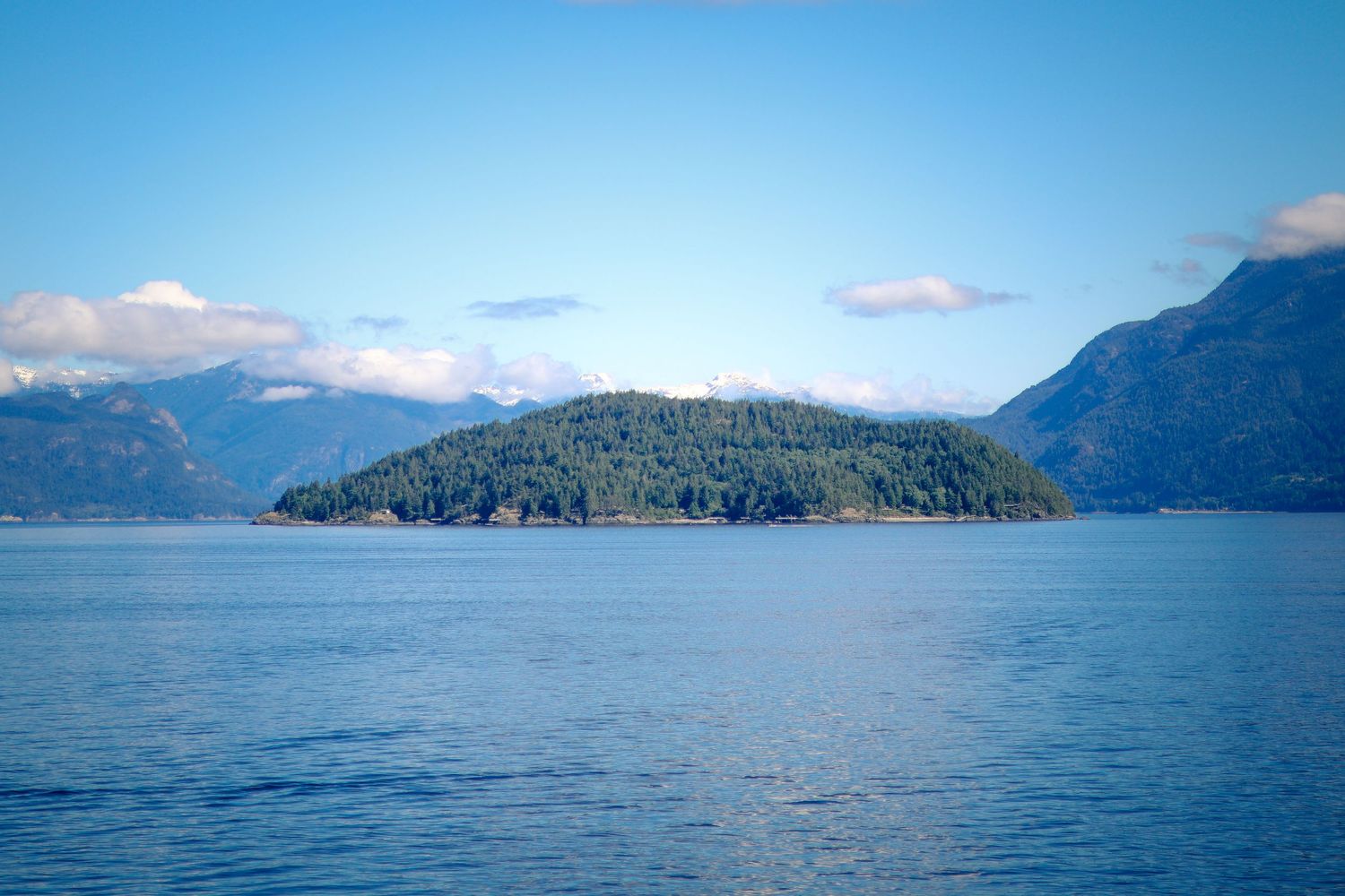 A forested island rises from calm blue waters surrounded by mountain ranges under a clear sky in the Pacific Northwest.
