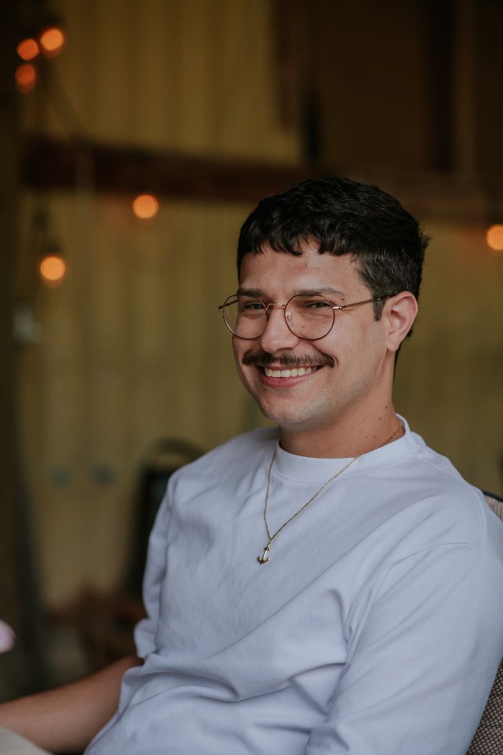 Smiling young man with glasses and mustache wearing a white sweatshirt and gold necklace seated in a warmly lit setting.