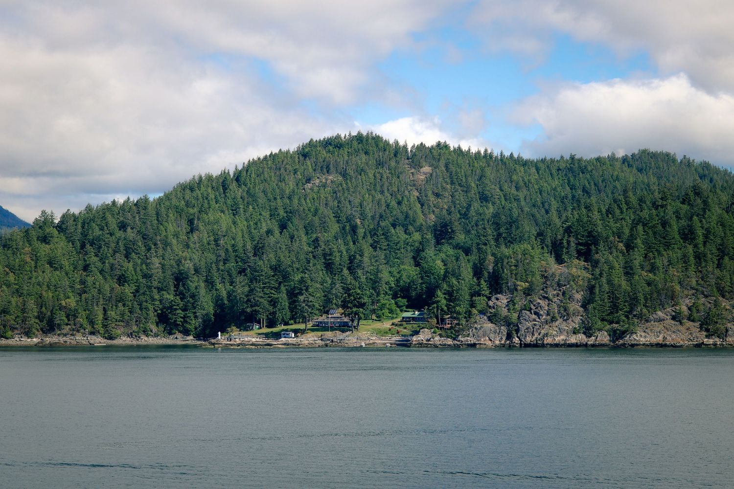 Lush green forested hillside rising above calm blue waters under a partly cloudy sky in a Pacific Northwest coastal landscape.