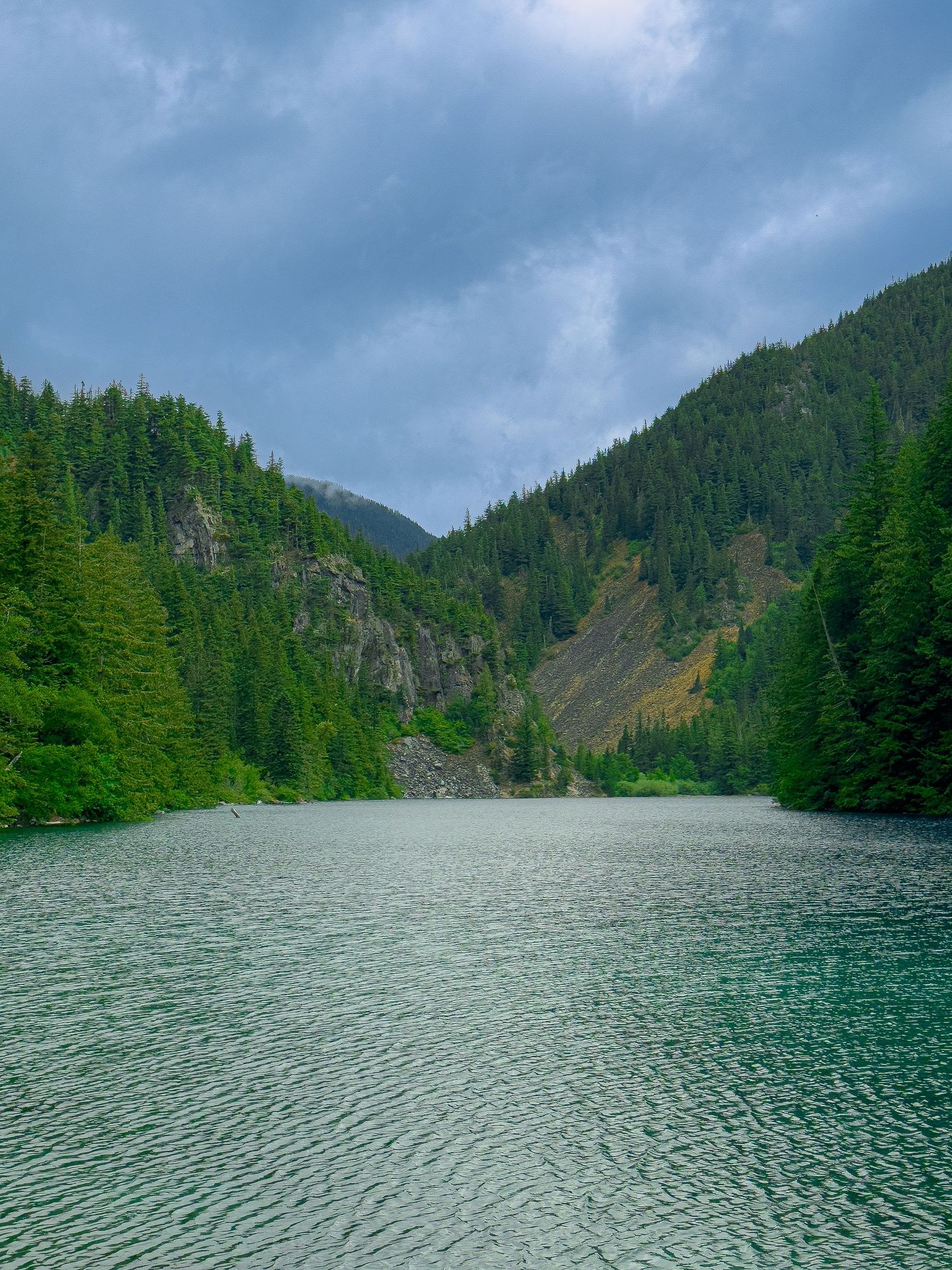 Serene turquoise mountain lake surrounded by dense evergreen forest and rocky cliffs under dramatic cloudy skies.