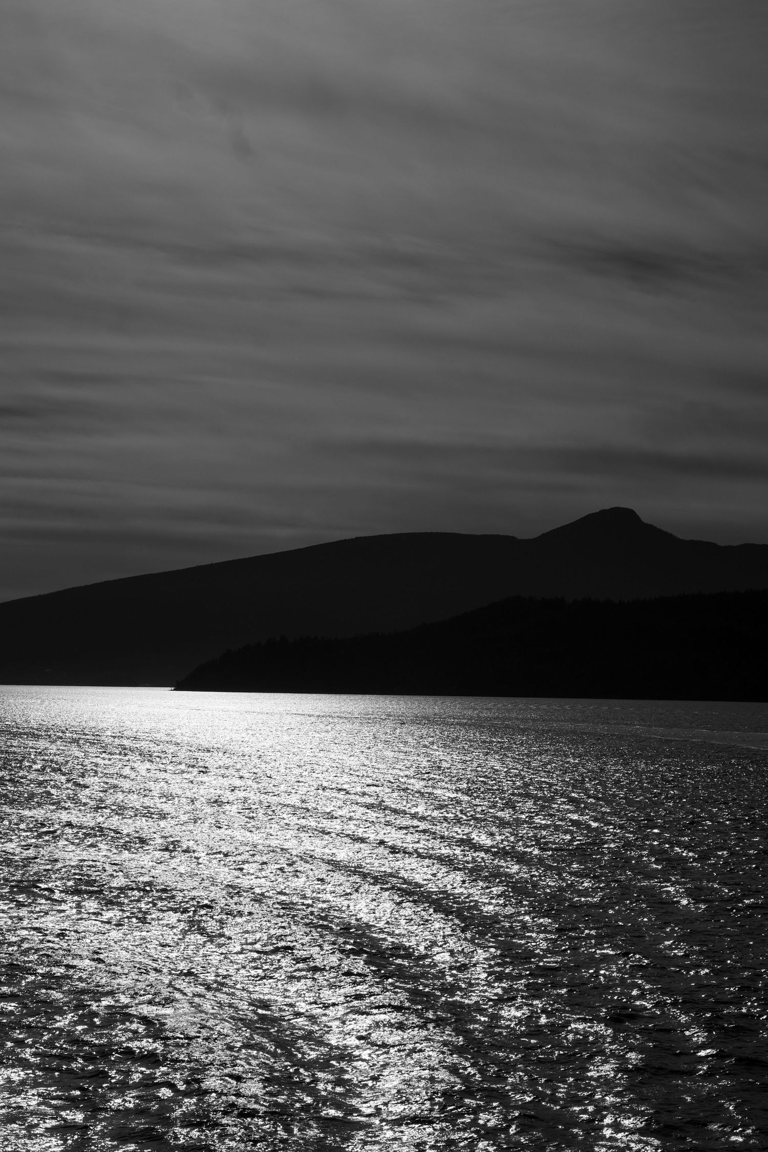 Black and white seascape with shimmering water reflections, mountain silhouette, and dramatic cloudy sky.