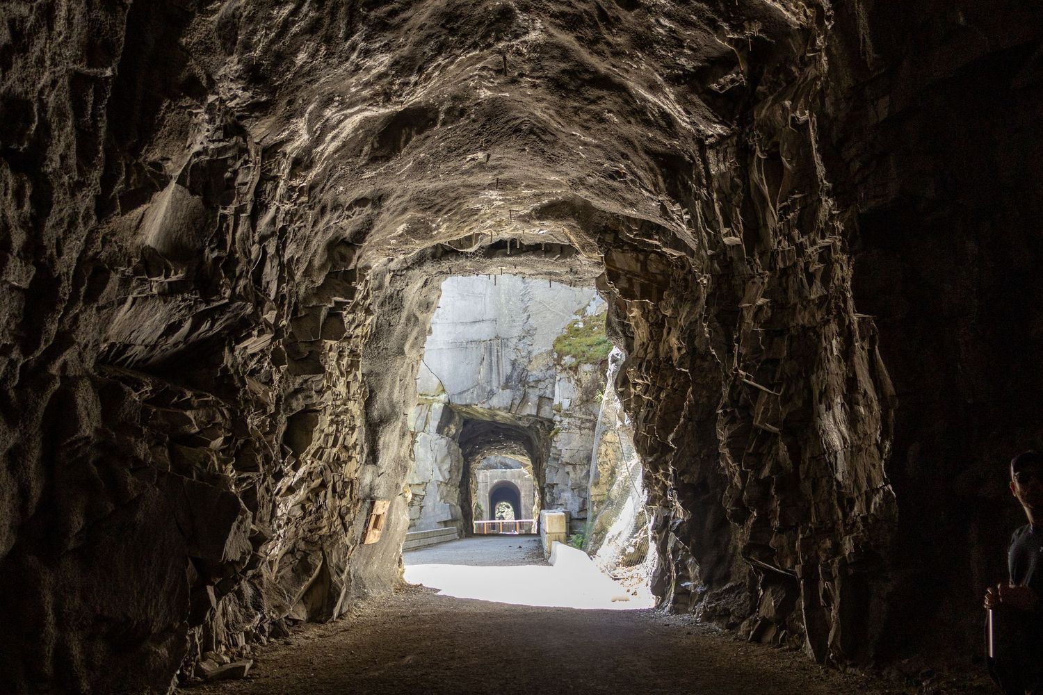 Rocky mountain tunnel with arched stone walls leading to a smaller lit tunnel opening, creating a dramatic perspective view.