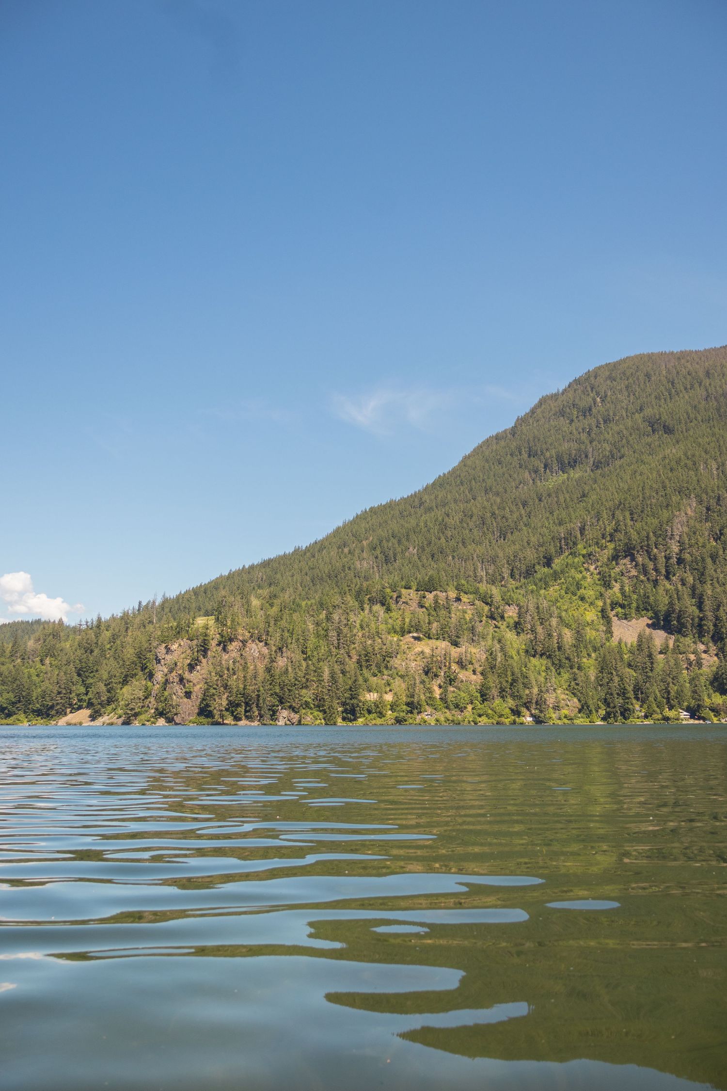 Serene mountain lake with calm green waters reflecting forested hillside under clear blue sky.